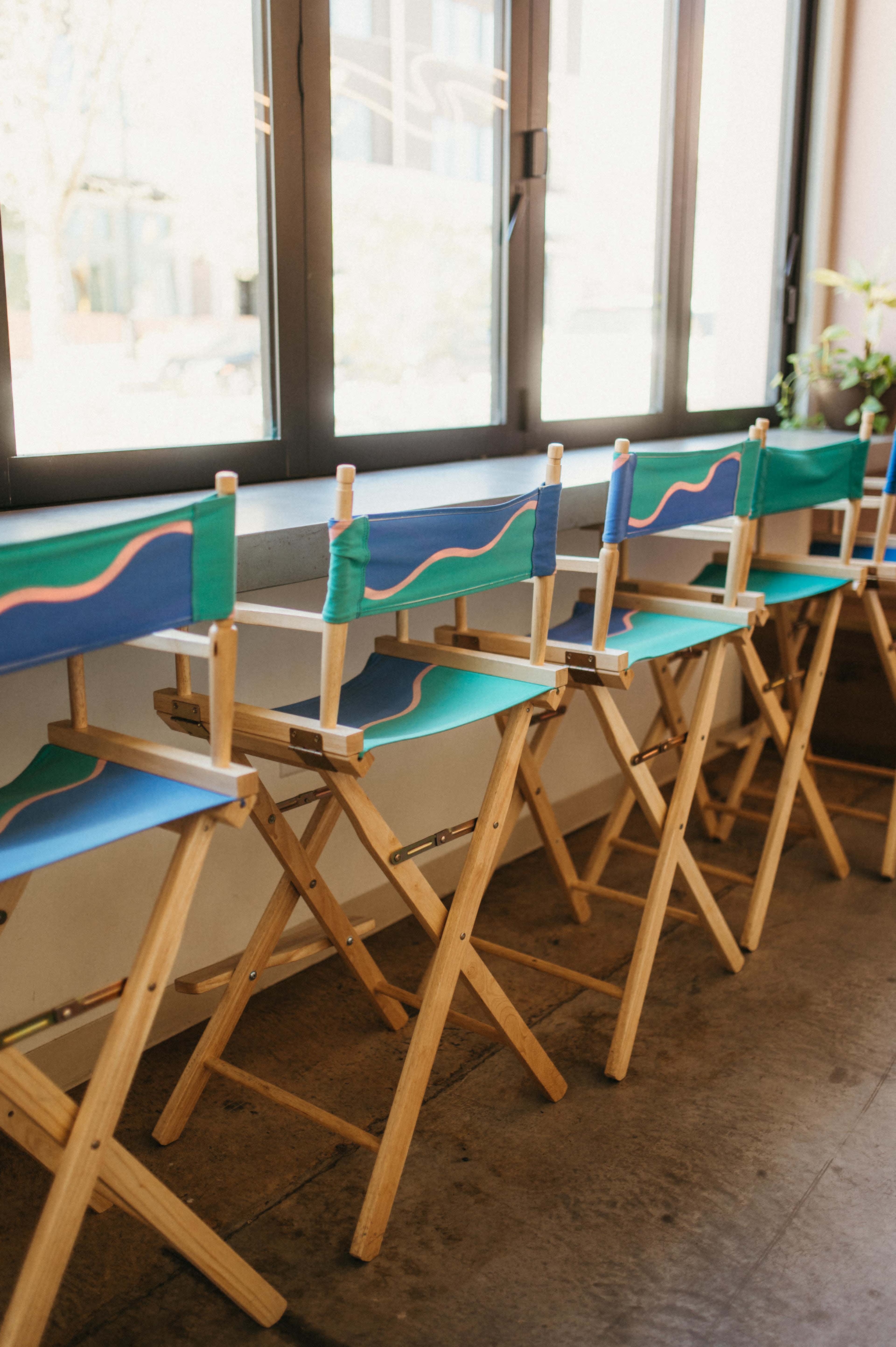 A row of wooden folding chairs with colorful fabric seat backs is lined up beside a large window.
