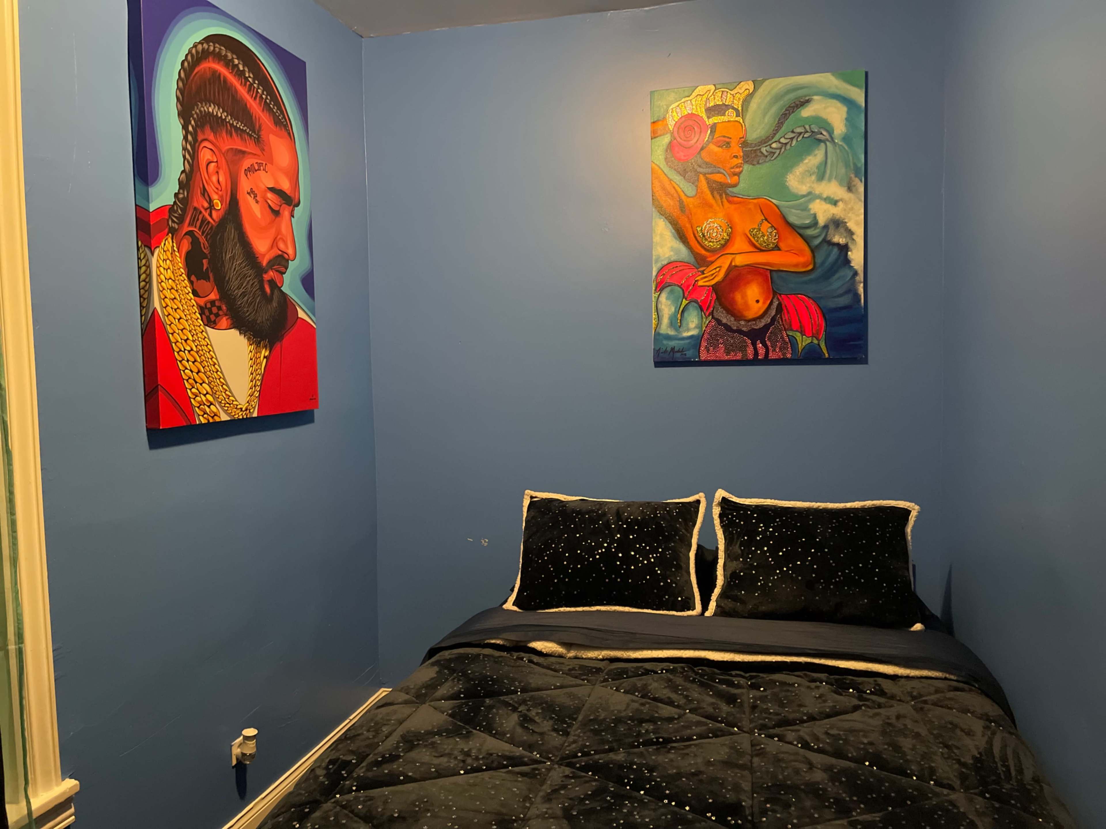 A bedroom with blue walls, a black bedspread adorned with decorative pillows, and two vibrant artworks featuring a man and a woman on the walls.