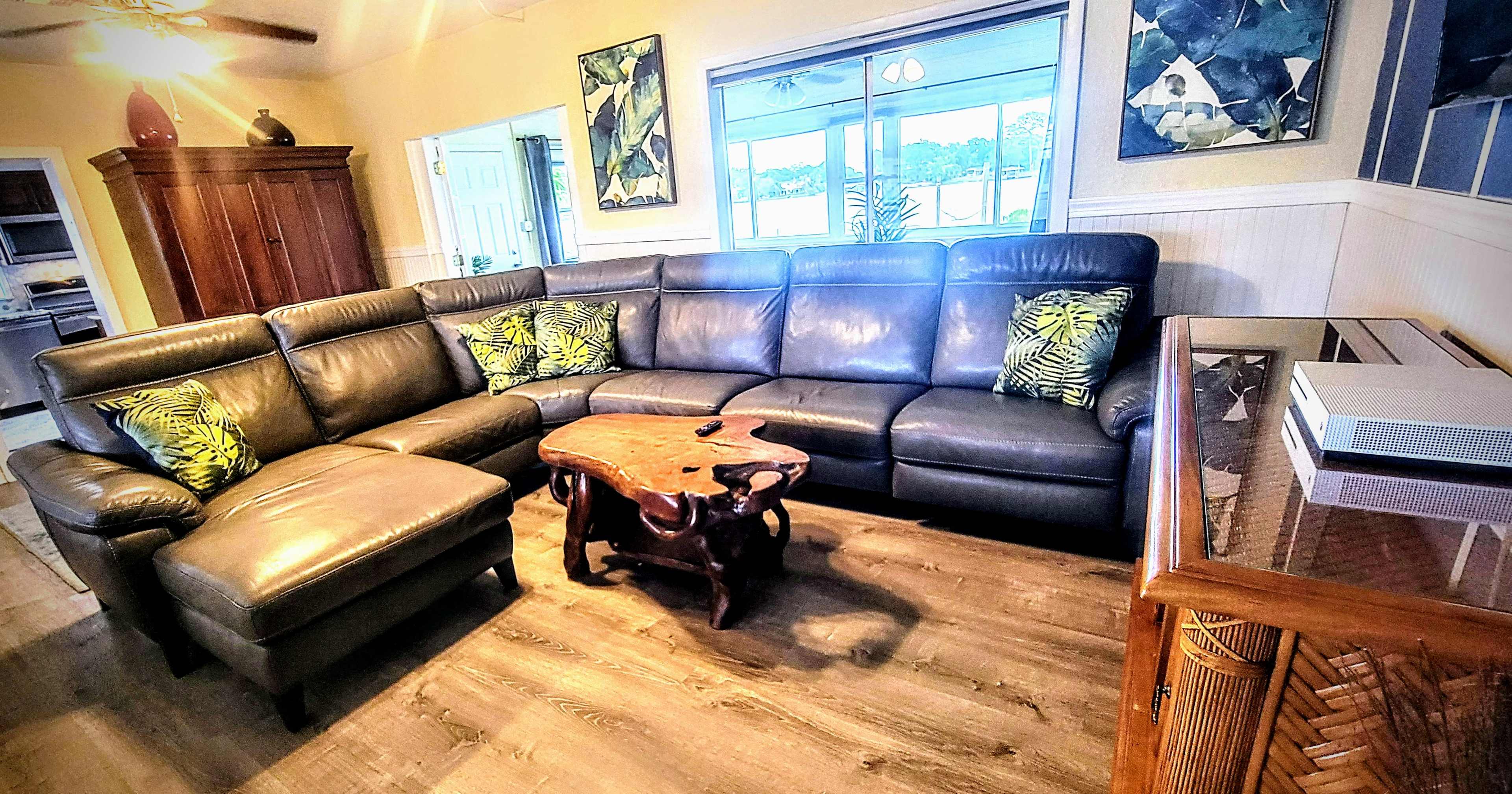 A living room with a dark leather sectional sofa, a wooden coffee table, and large windows allowing light to enter.