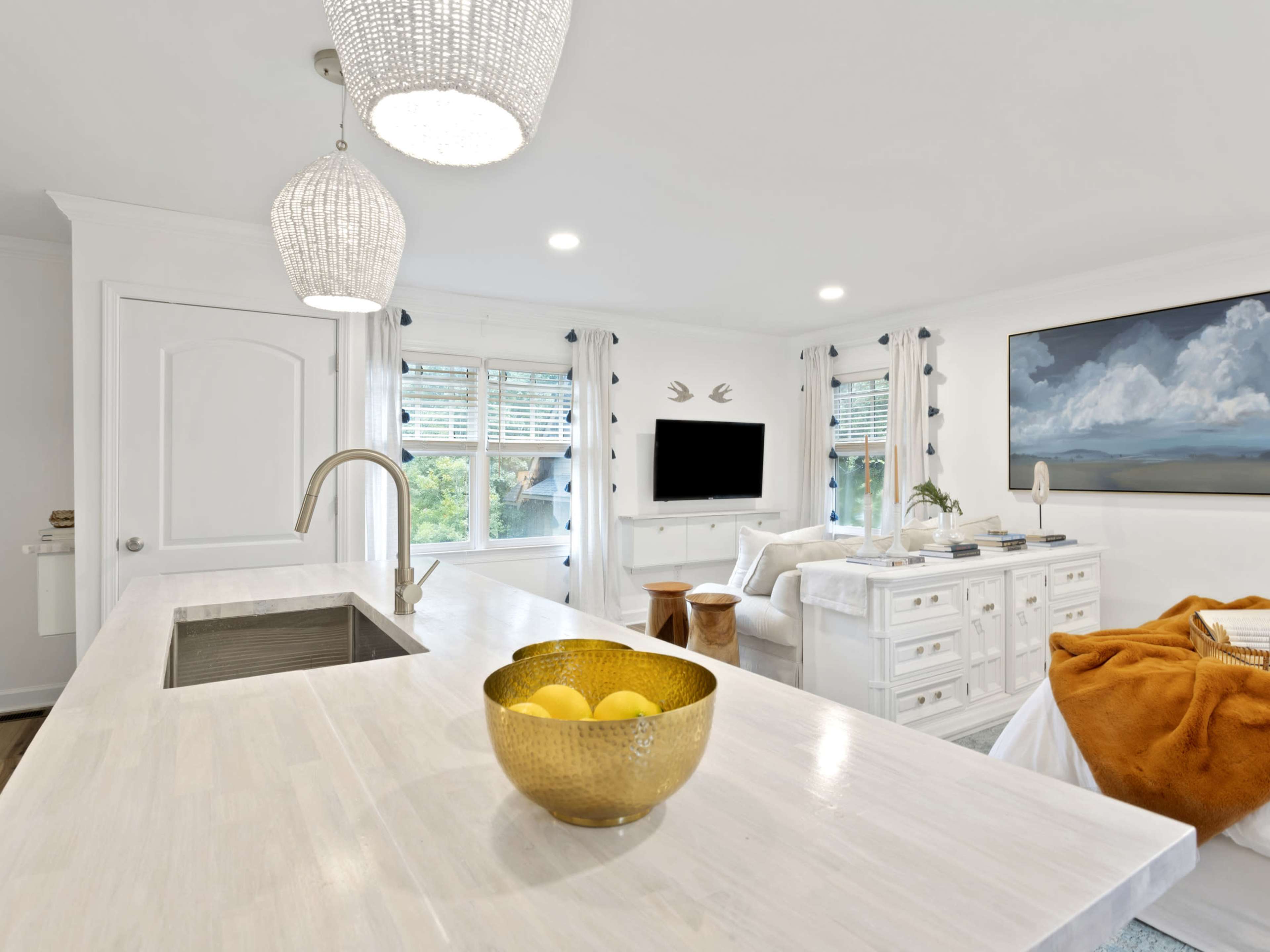 A modern white kitchen features a spacious island with a sink and a bowl of lemons, set against a living area with a television and artwork.