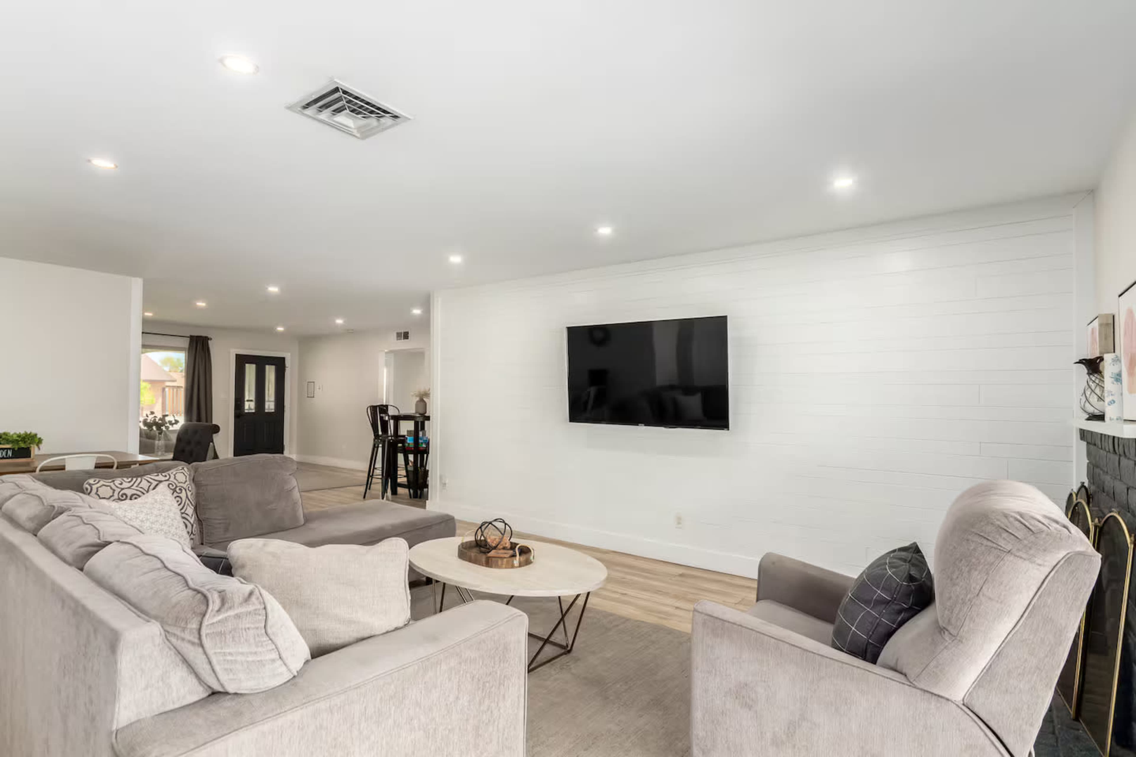 The image shows a modern living room featuring a light-colored sofa arrangement, a round coffee table, a mounted television on a white wall, and hardwood flooring.
