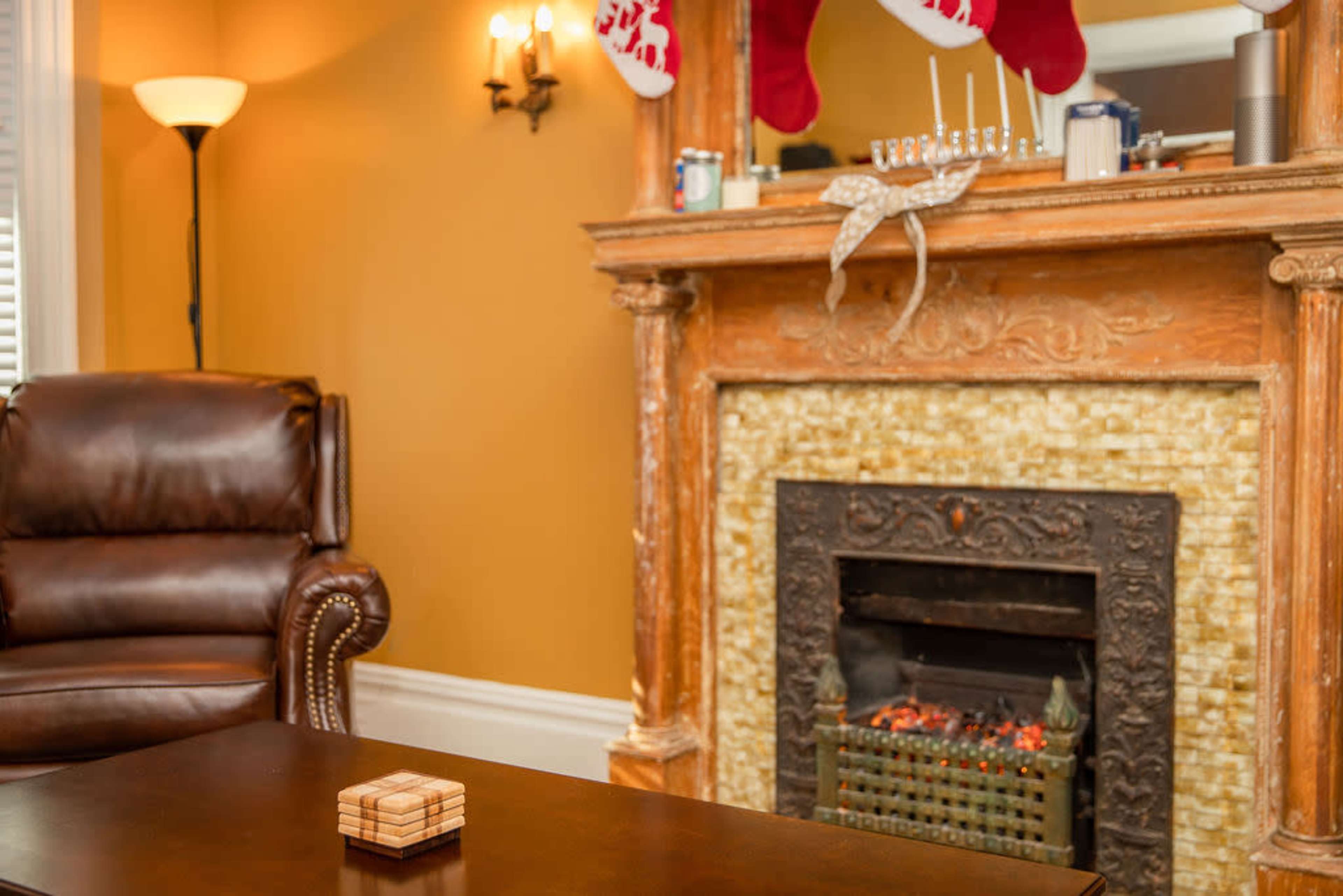 The image shows a cozy living room with a brown leather armchair, a decorative fireplace with a stone facade, and stockings hung on the mantel.
