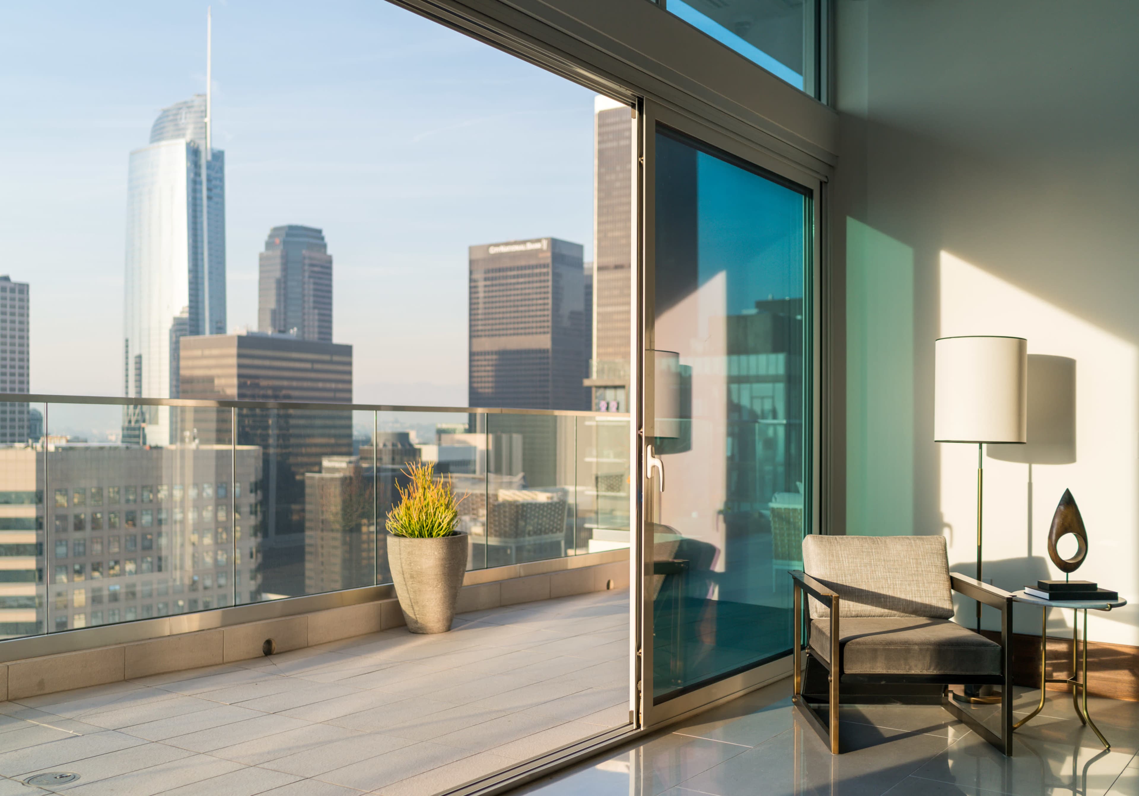 A modern room with large glass doors opens to a balcony overlooking a city skyline, featuring a potted plant and a small chair.