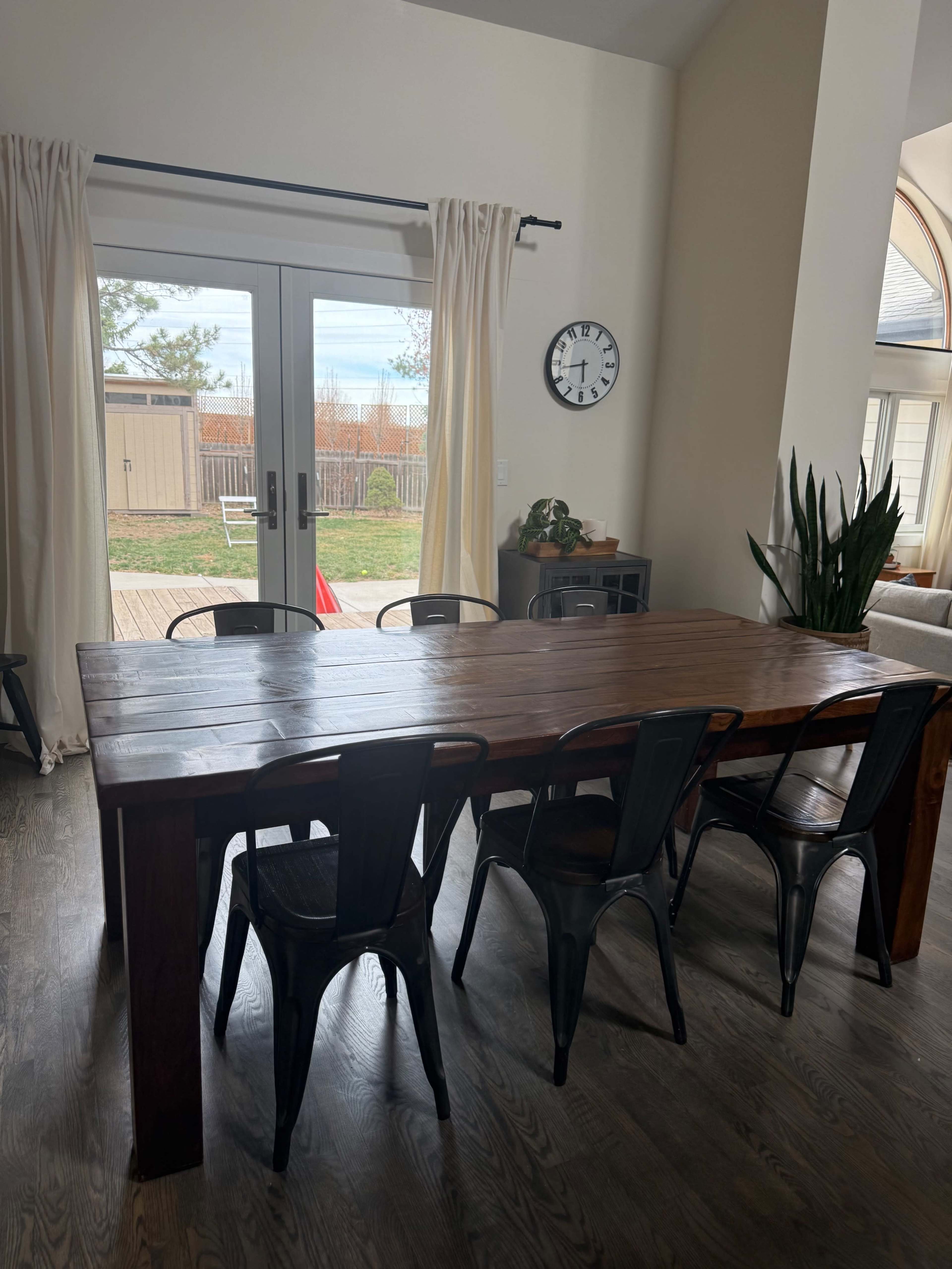 The image shows a wooden dining table surrounded by black metal chairs in a room with large windows and sliding glass doors.
