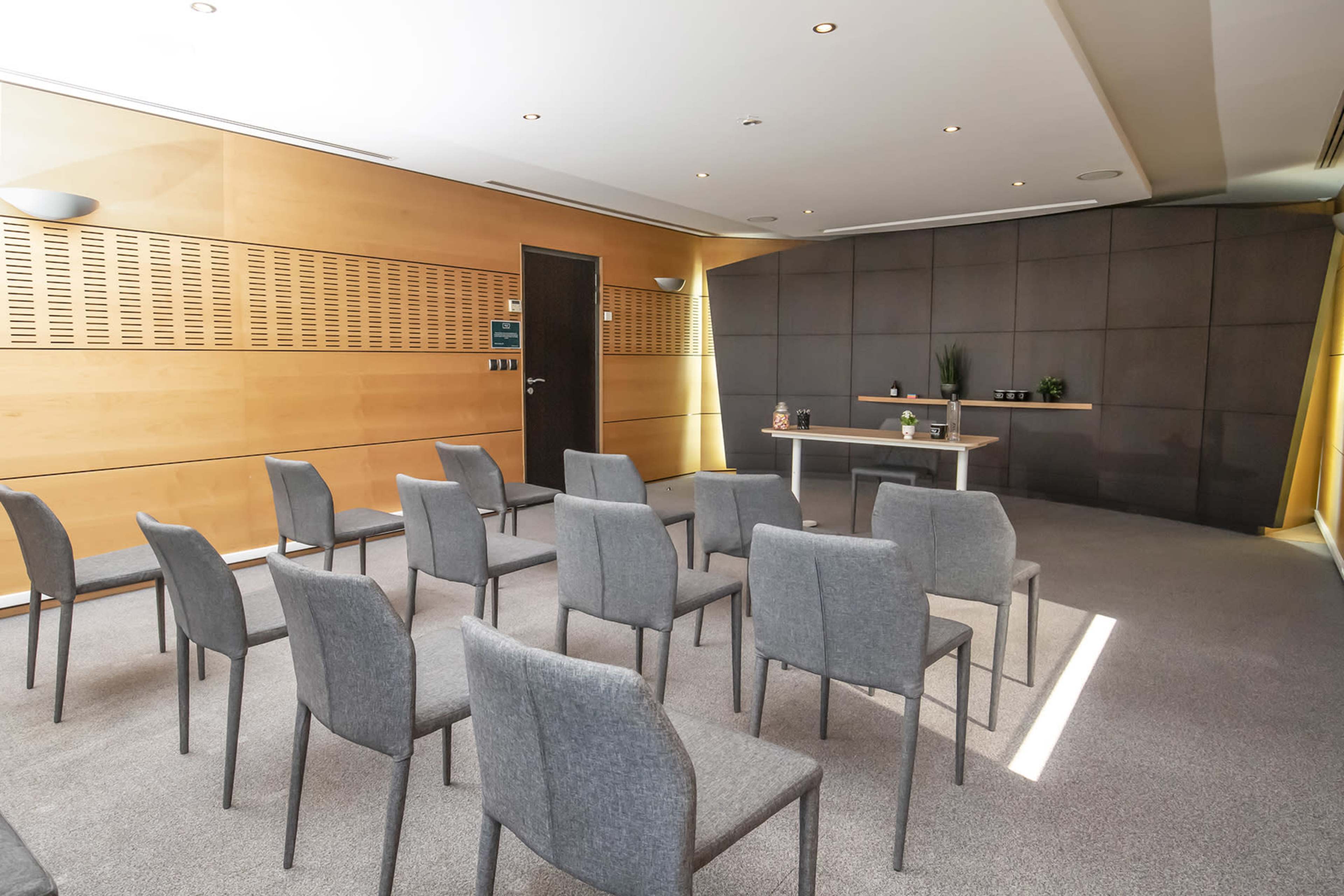A small presentation room is set up with gray chairs arranged in rows facing a central table against a backdrop of wooden and gray walls.