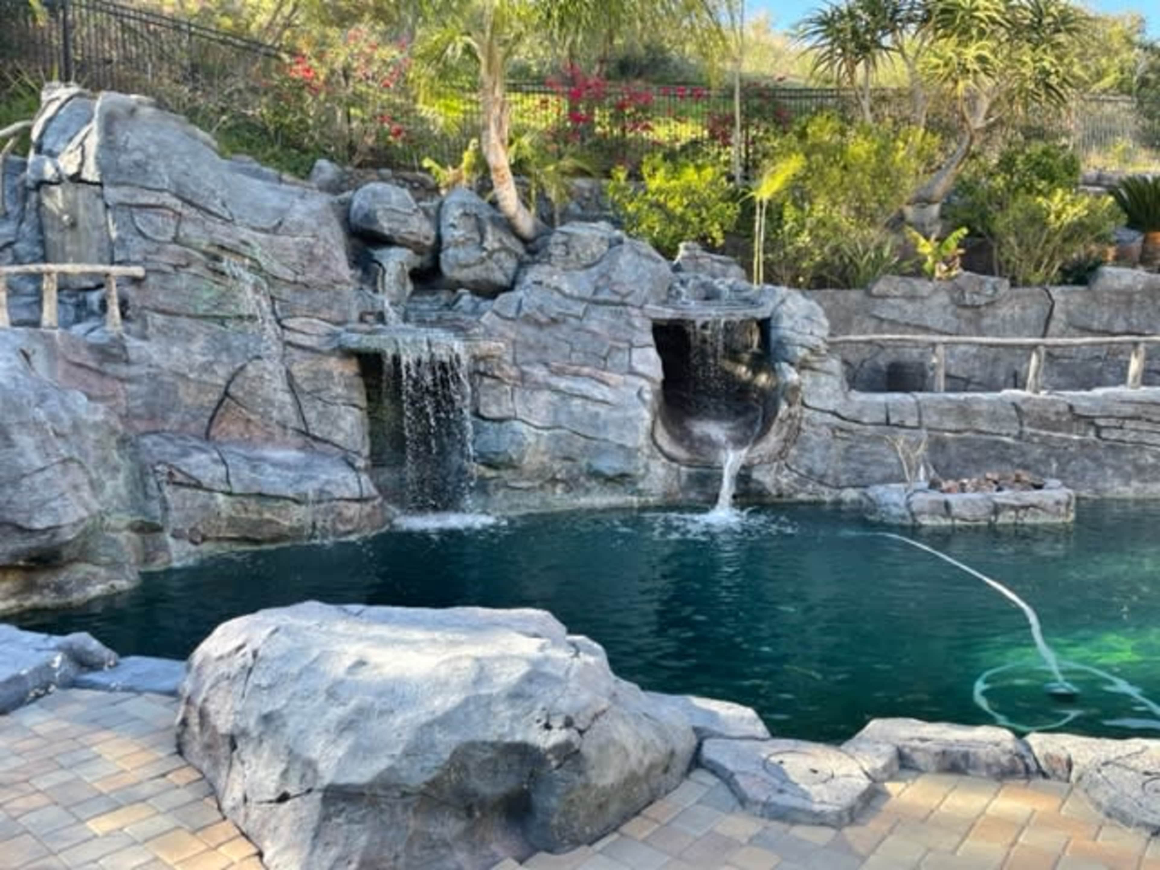 The image shows a landscaped pool area with rock formations, a small waterfall, and surrounding greenery.