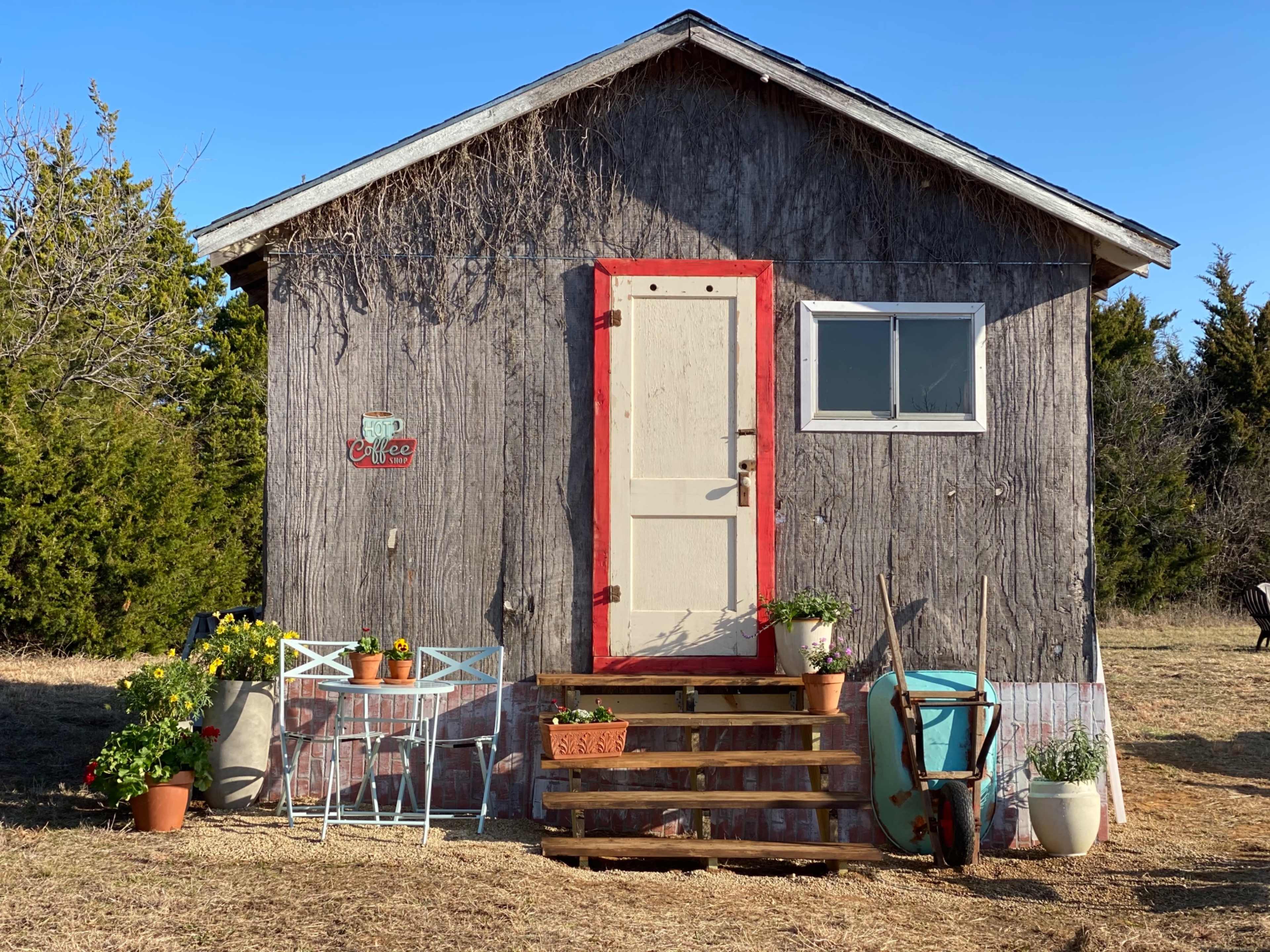 A small wooden cabin with a red door and two white chairs, adorned with potted plants, sits in an open field.