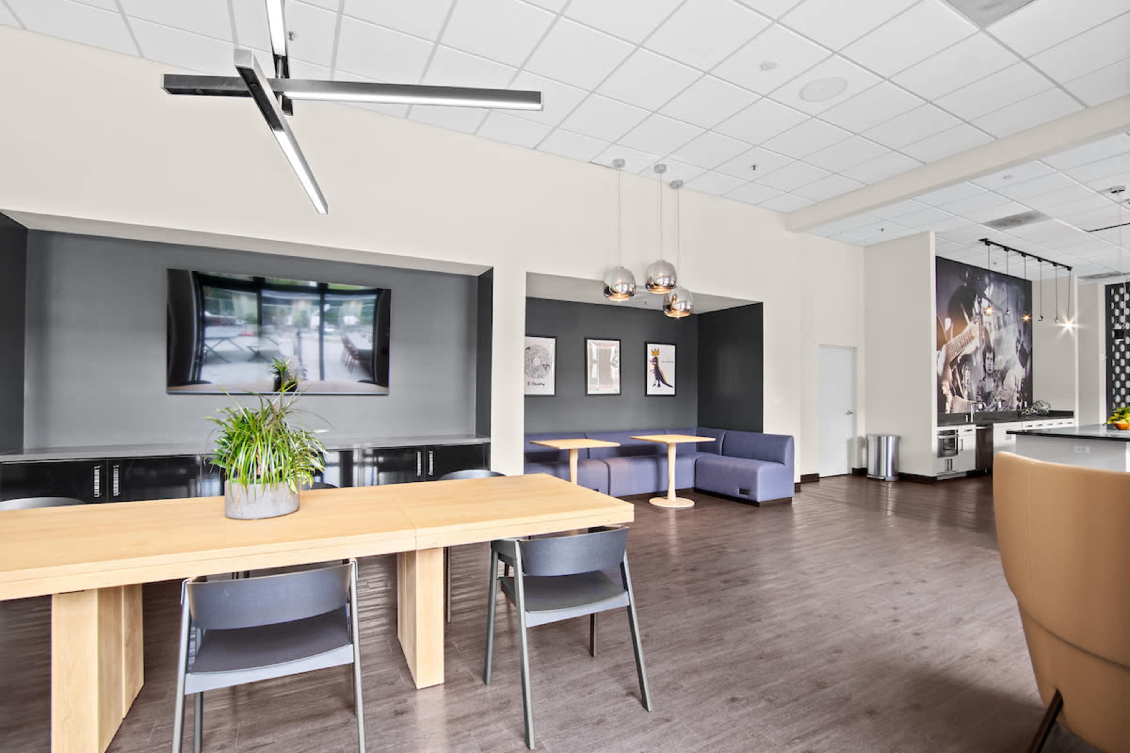 The image shows a modern lounge area featuring a wooden dining table, grey chairs, a large television on the wall, and a kitchenette in the background.