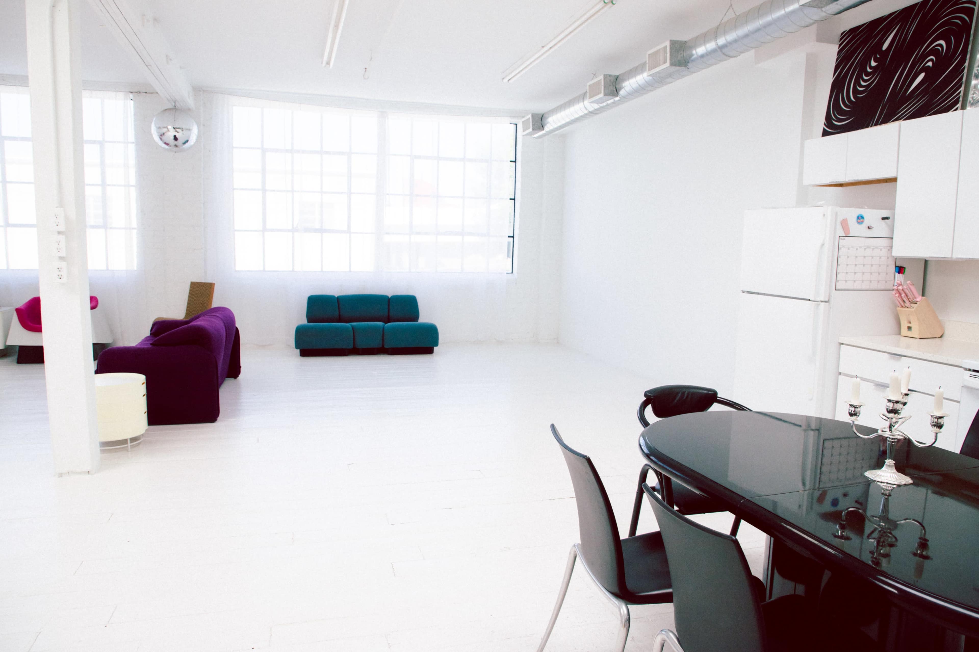 A spacious, bright room with white walls and floors, featuring a black dining table and chairs, a kitchen area, and two colorful sofas in the living space.