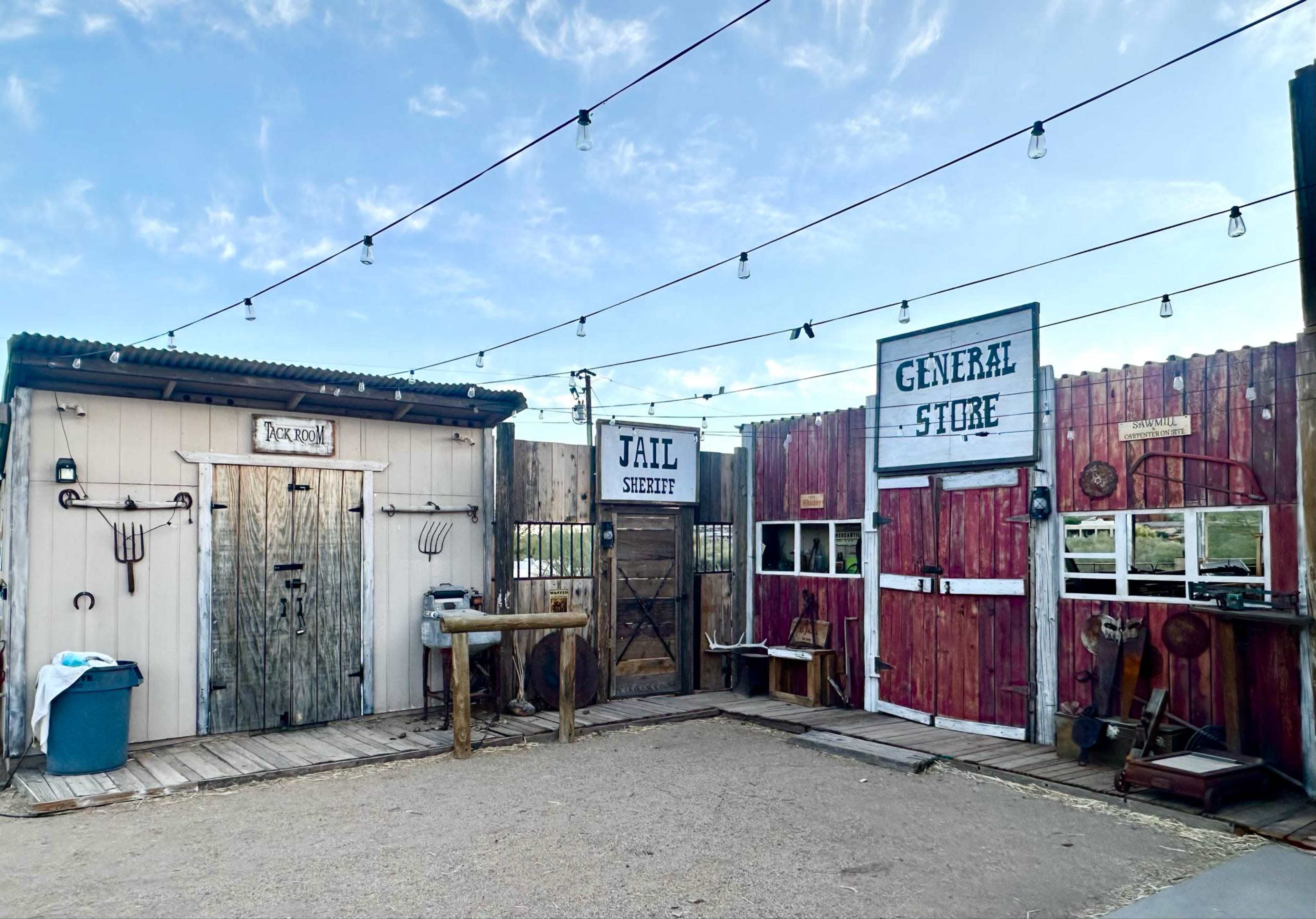 The image shows a western-themed setting featuring a "Jail," a "General Store," and a "Saloon," with wooden structures and string lights hanging above.