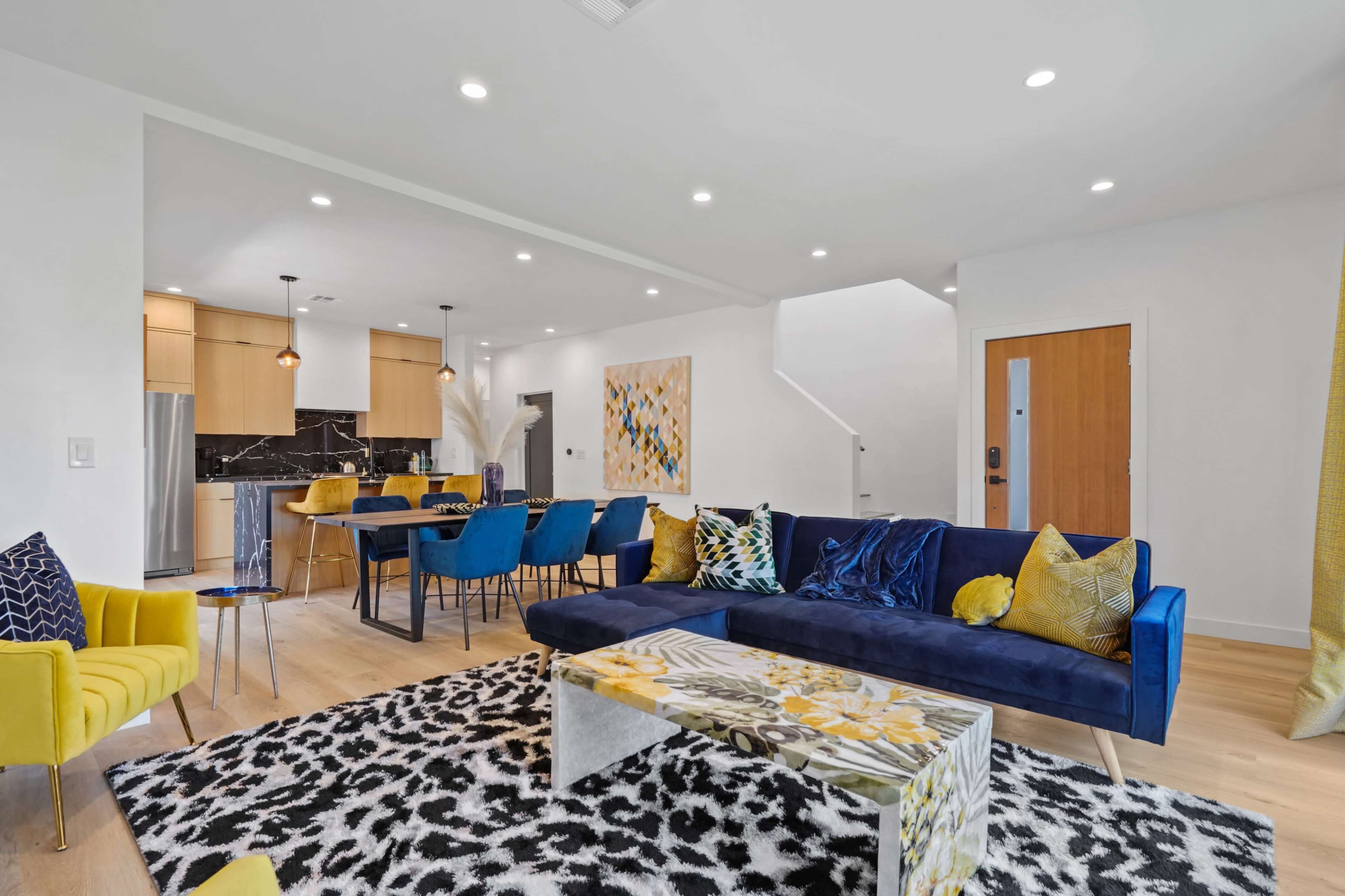 A modern living and dining area featuring a navy blue couch, colorful chairs, a patterned rug, and a contemporary kitchen in the background.
