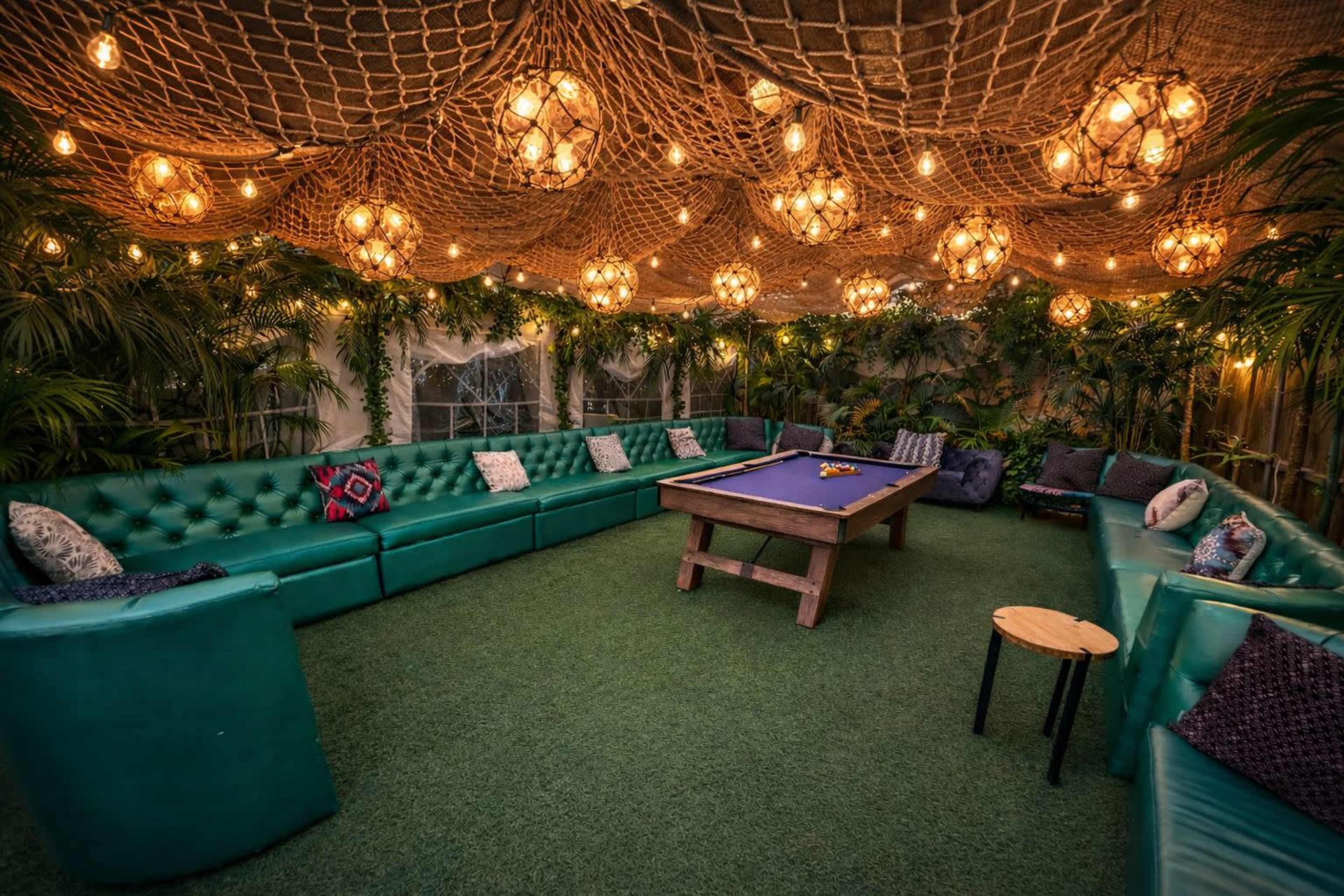 The image shows a cozy lounge area with green upholstered seating, a pool table in the center, and decorative string lights hanging from a netted ceiling.