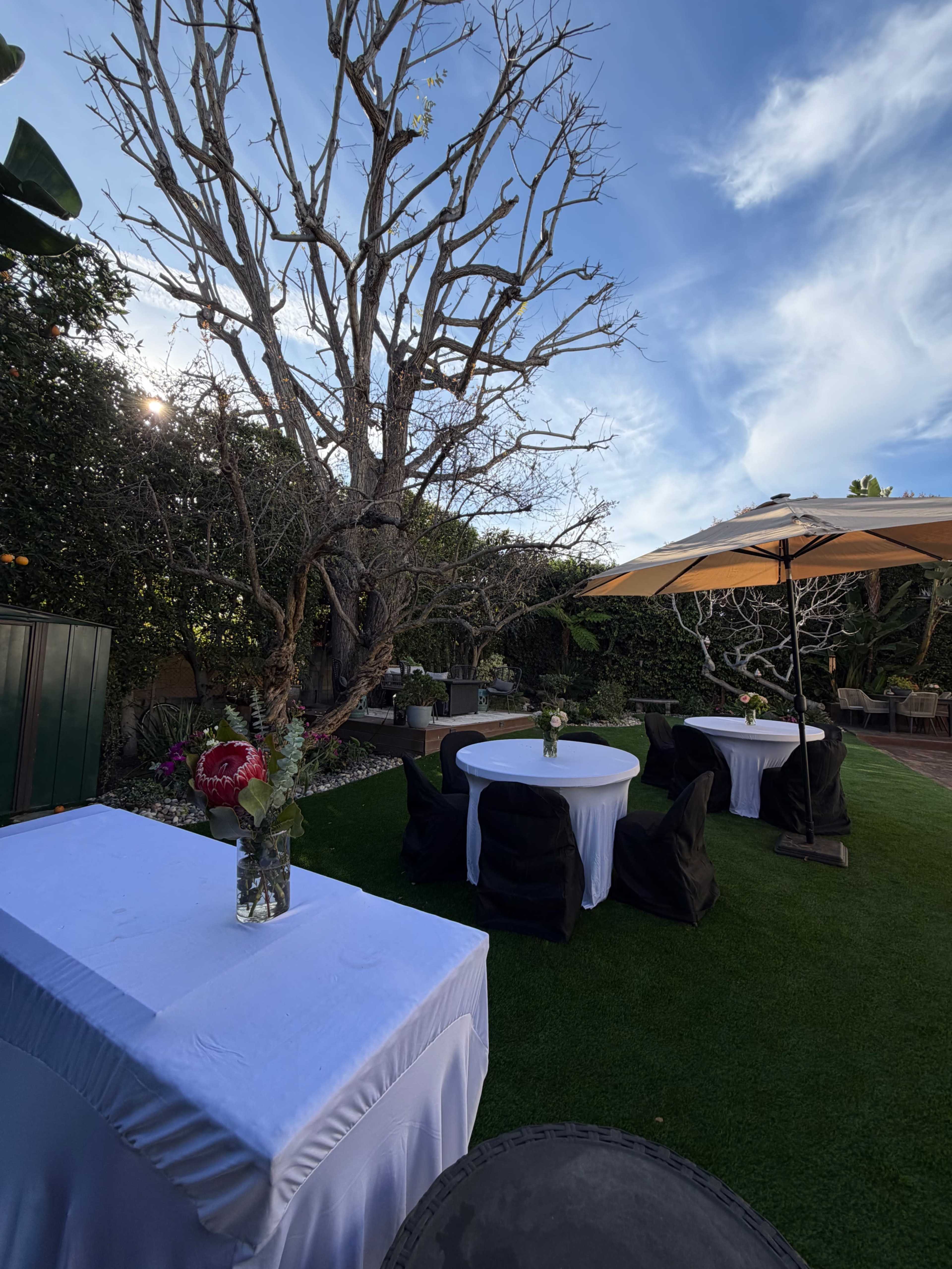 The image shows a garden setting with three round tables covered in white tablecloths and black chair covers, surrounded by trees and a large umbrella.