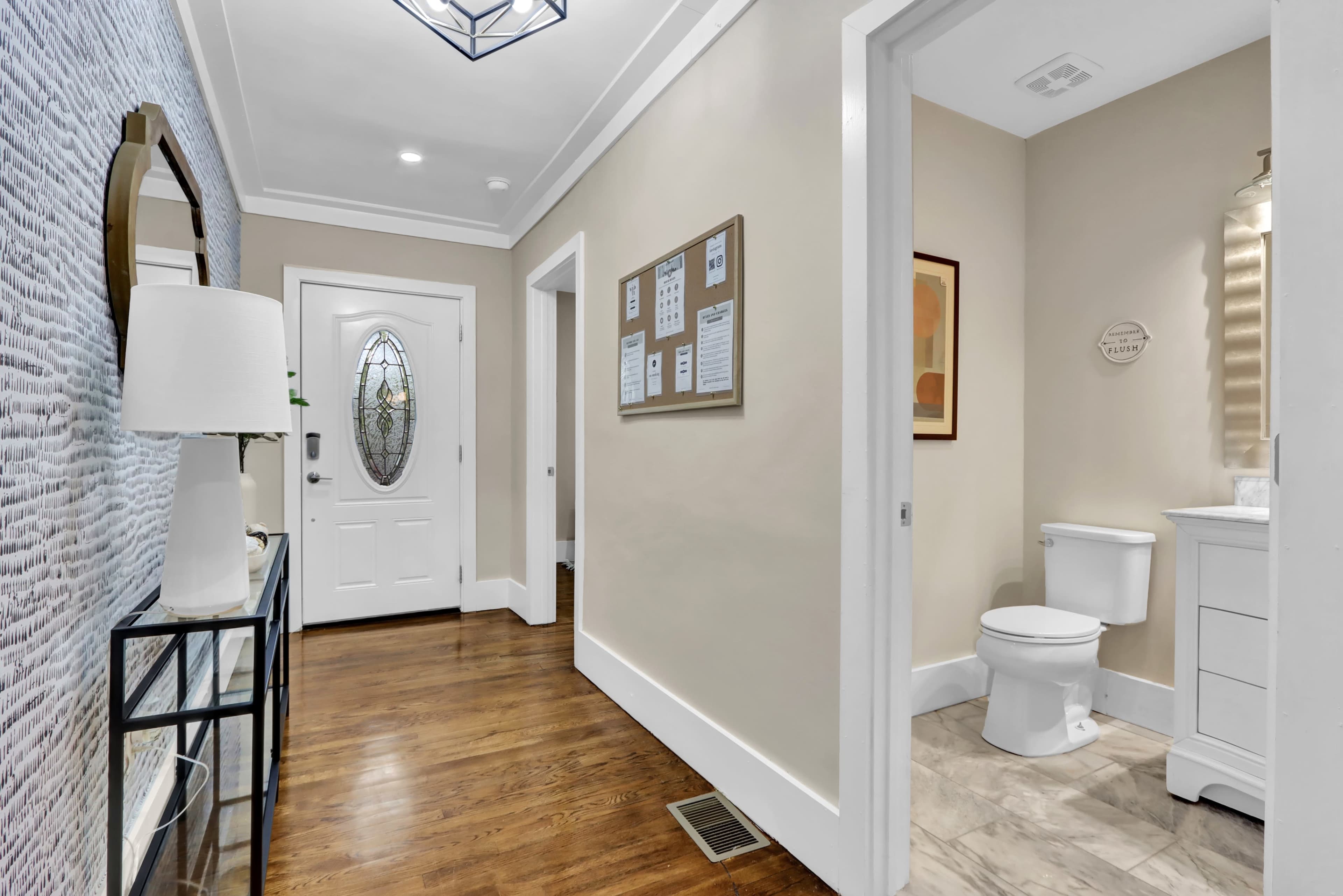 A hallway with a decorative mirror and white wall decor, leading to a bathroom on the right and a front door at the end.