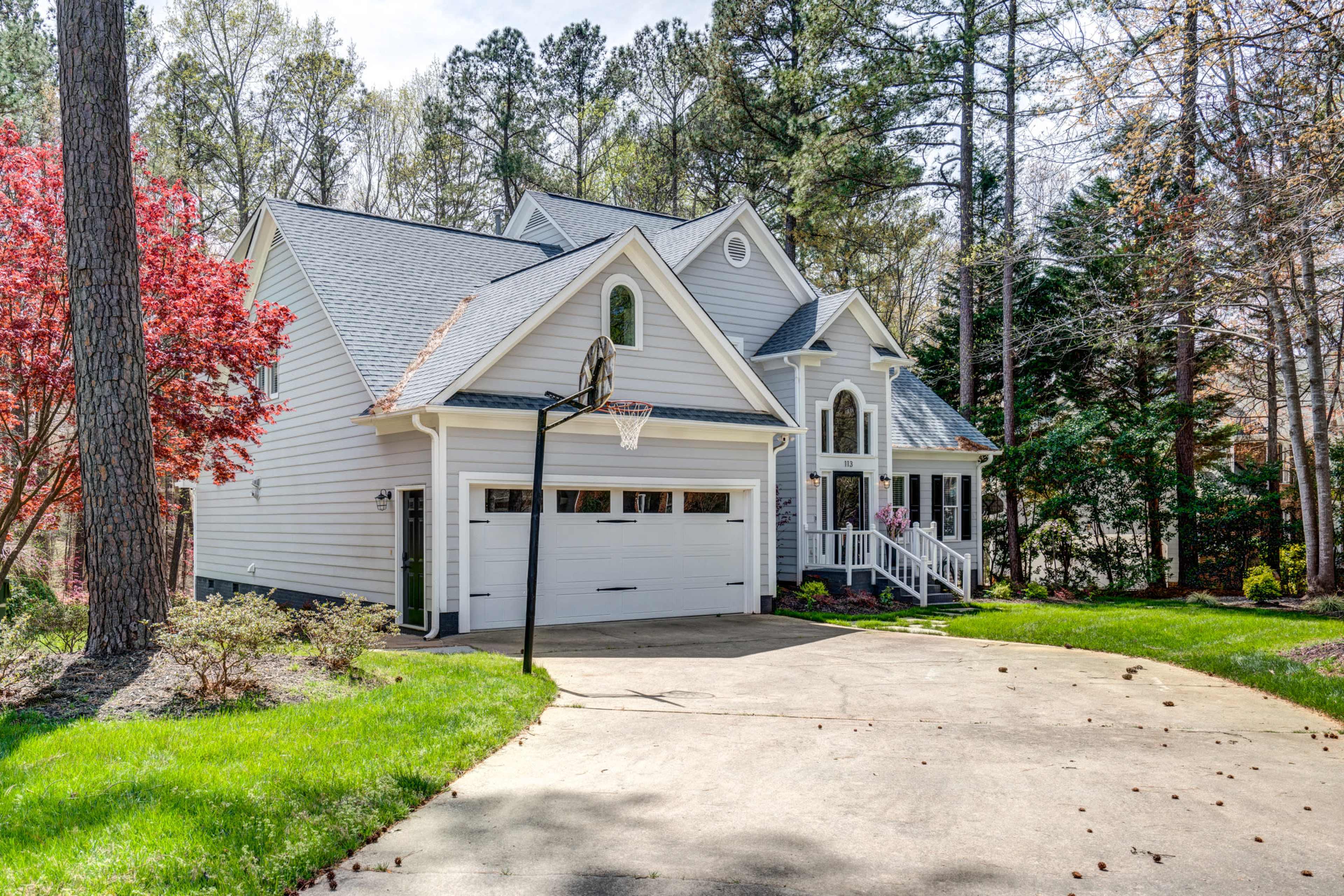 A large, light-colored house with a steep roof, a basketball hoop in the driveway, and surrounded by trees and green grass.