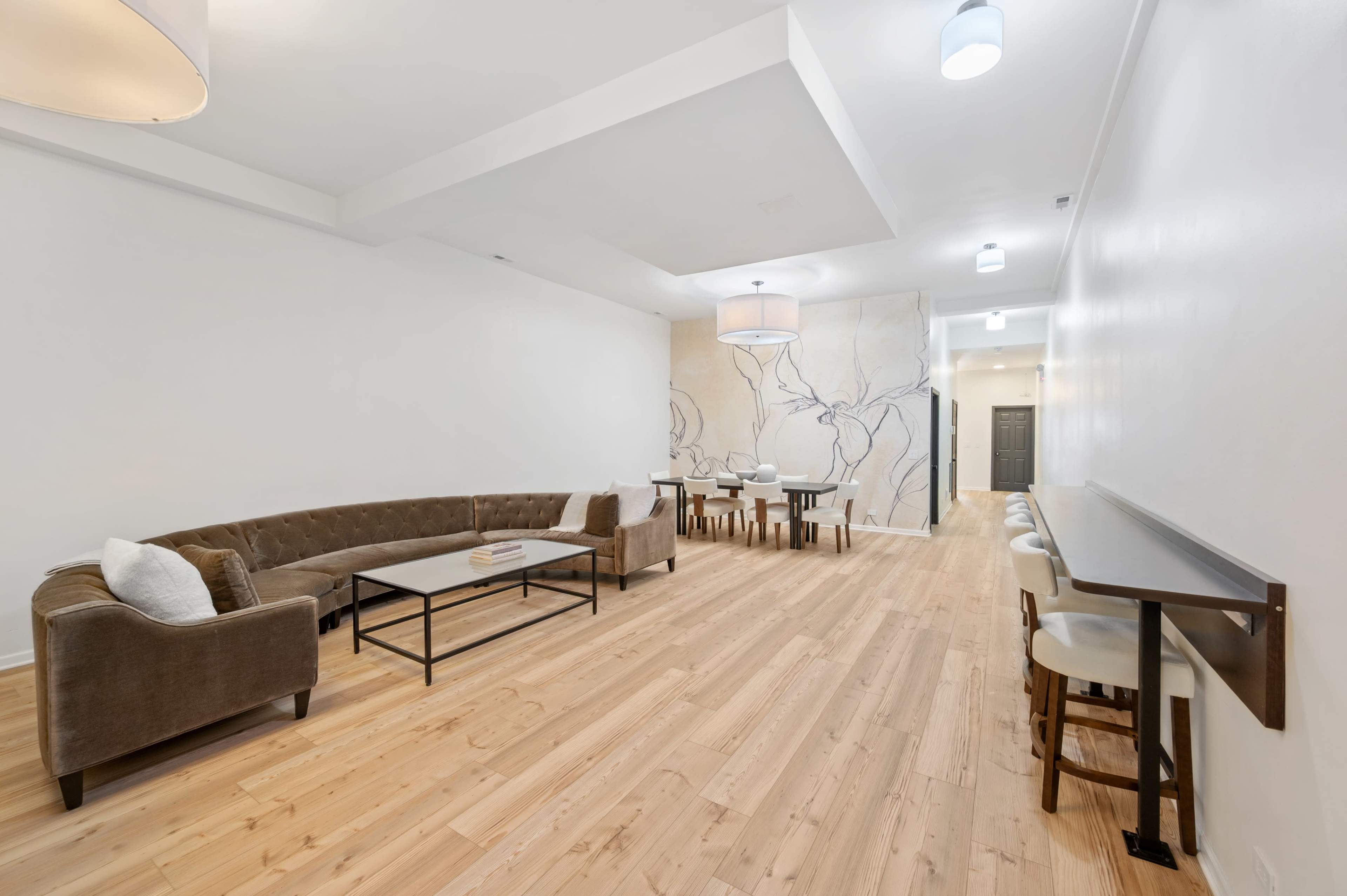 The image shows a spacious interior with light wood flooring, a large sofa, a dining area with several chairs and tables, and a minimalist design featuring a floral mural on one wall.