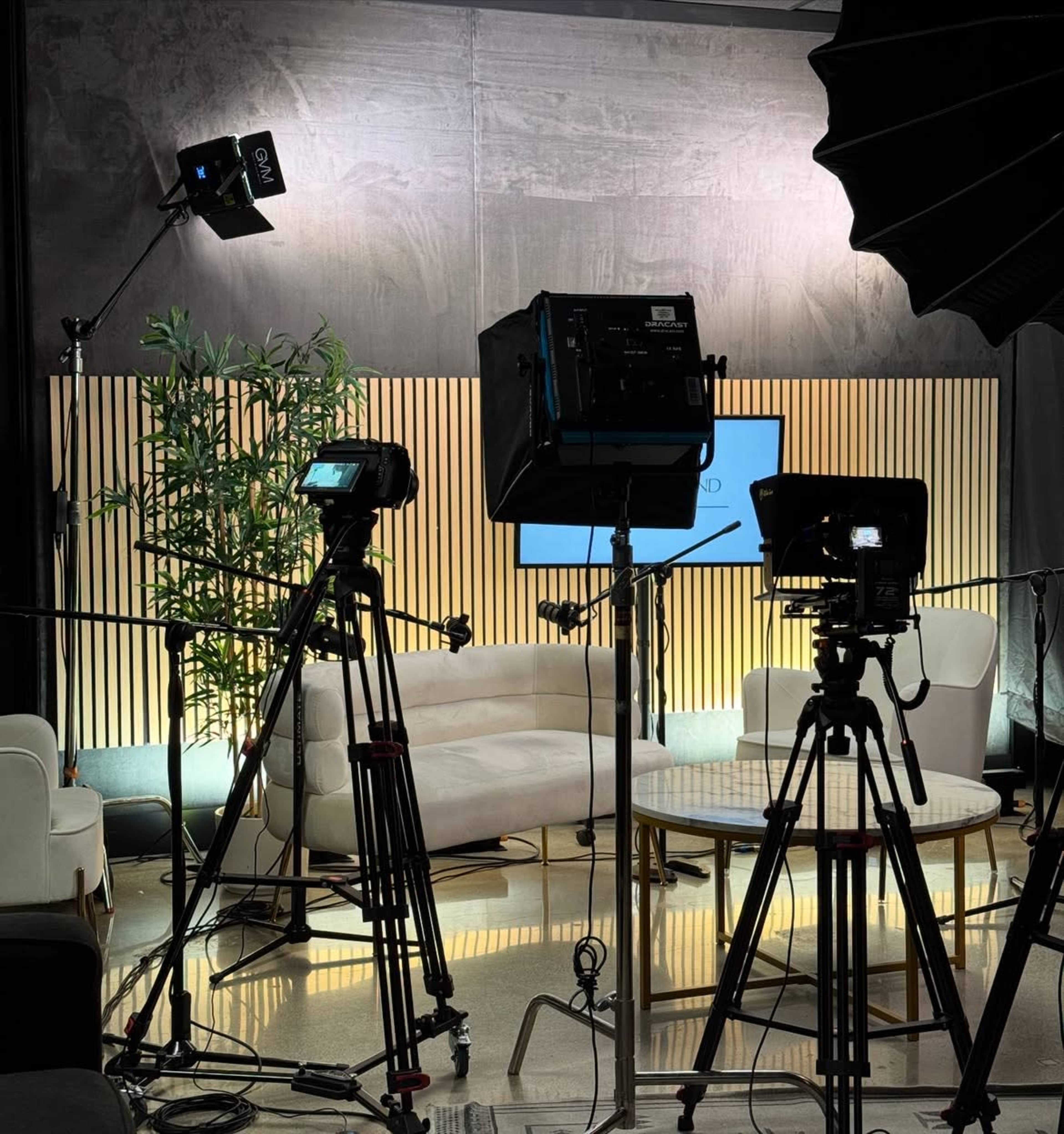 The image shows a well-lit studio setup with cameras, lighting equipment, and a sofa against a backdrop of wooden slats.