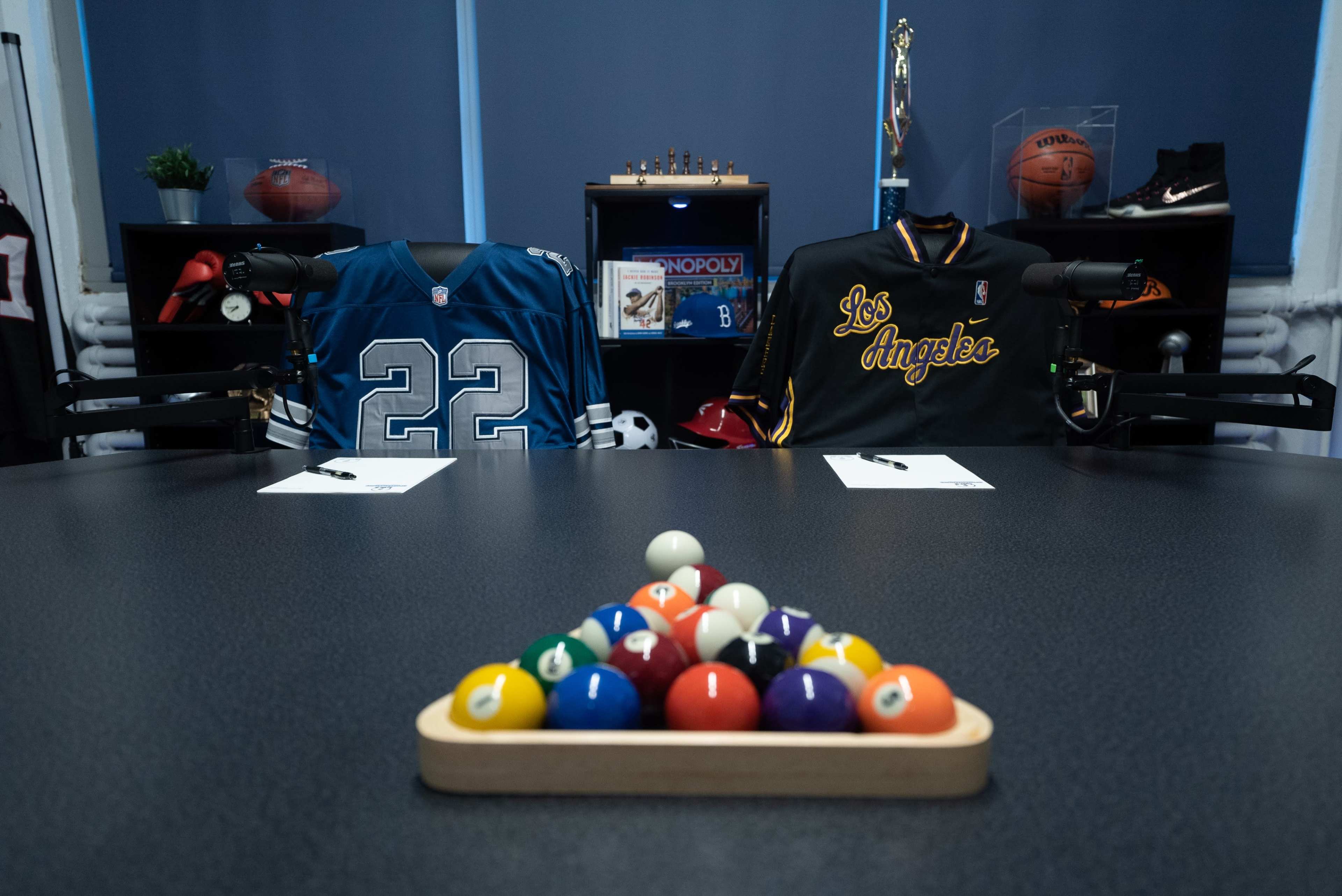 A table set for discussion with two sports jerseys, a set of billiard balls arranged in a triangle, and notepads on either side, in a room decorated with sports memorabilia.