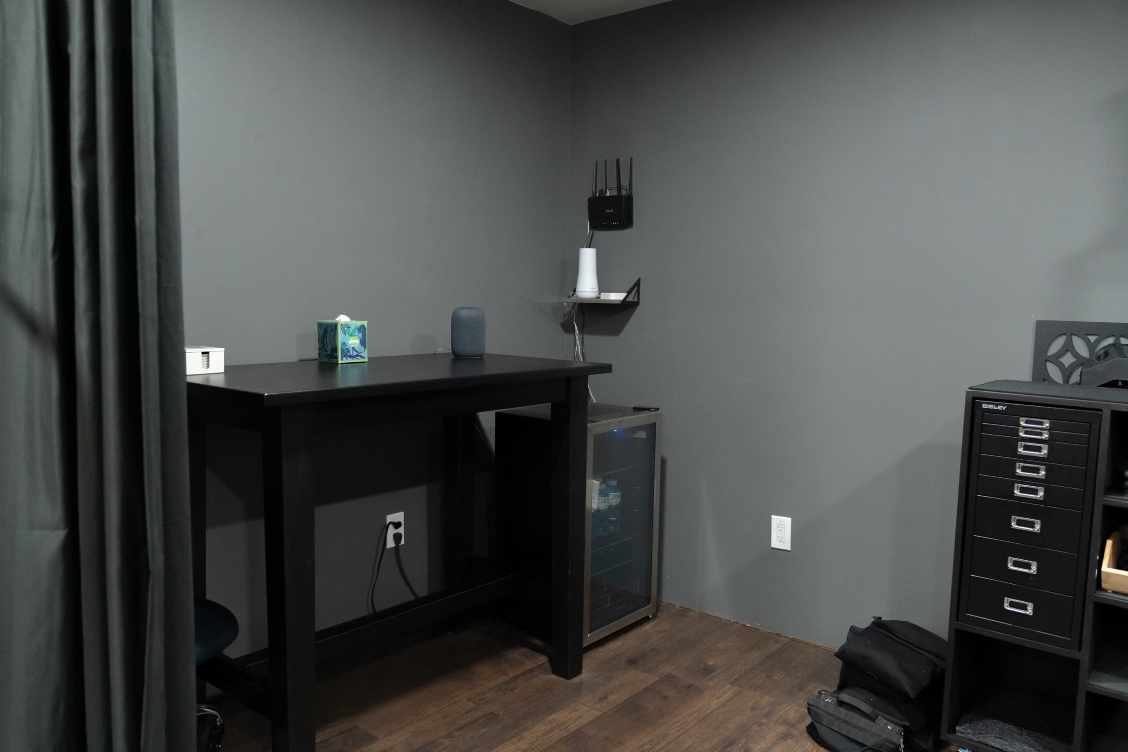 The image shows a corner workspace with a black desk, a small wall-mounted shelf, and a refrigerator beside a filing cabinet against a gray wall.