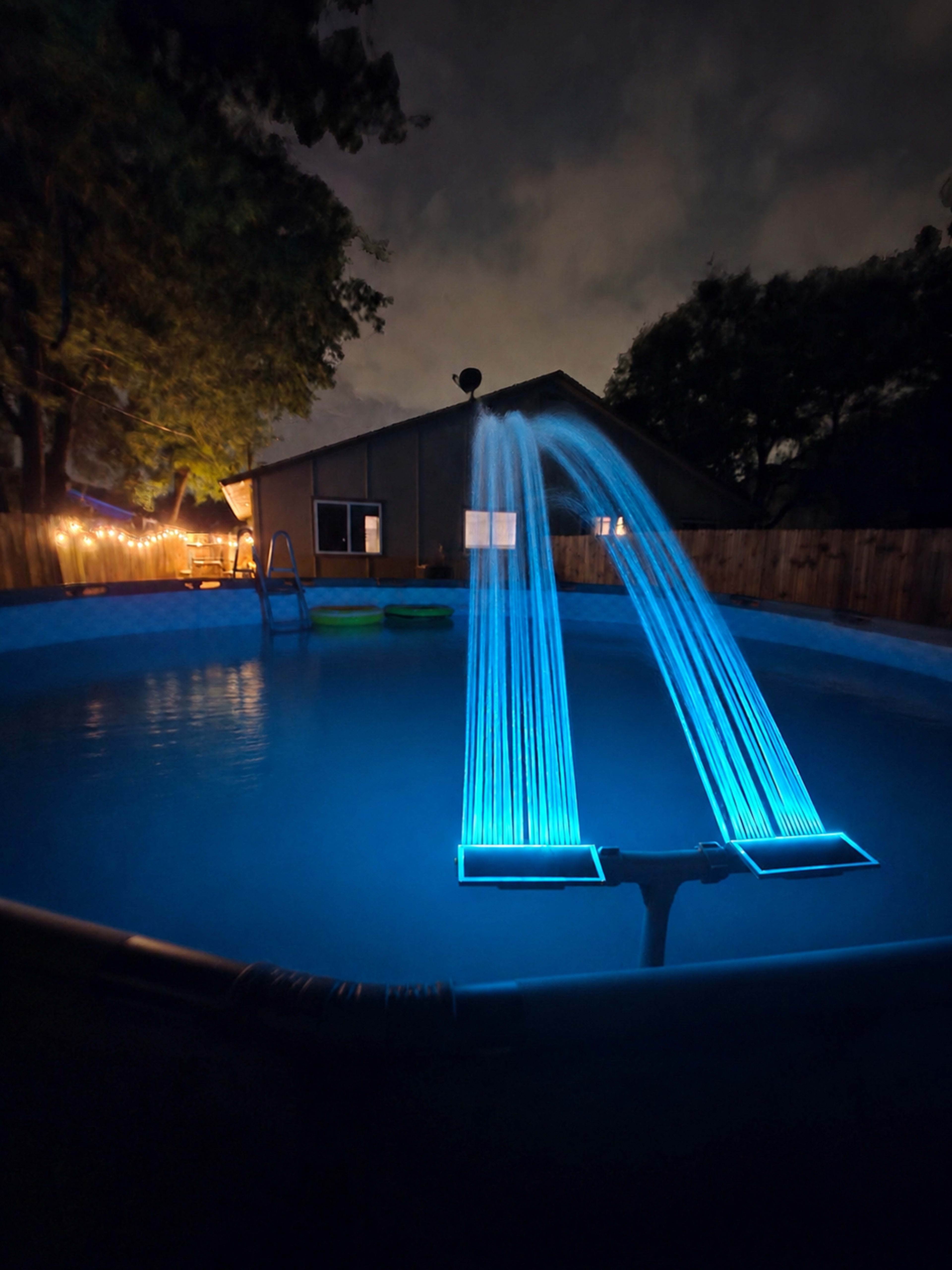 A lighted water feature creates arcs of blue water in a swimming pool surrounded by a fenced backyard at night.