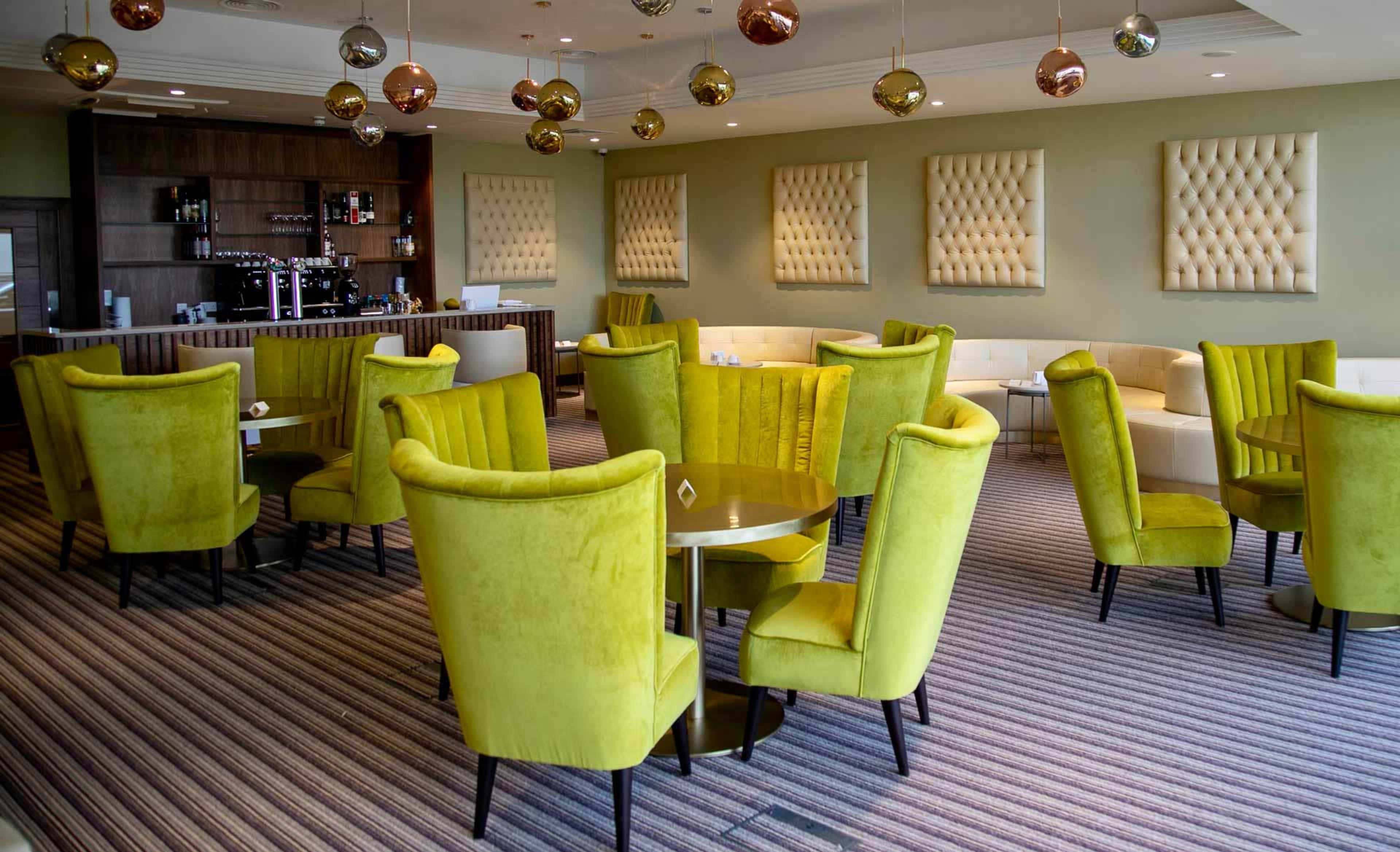 A spacious café with green upholstered chairs arranged around small round tables, with a bar area in the background.