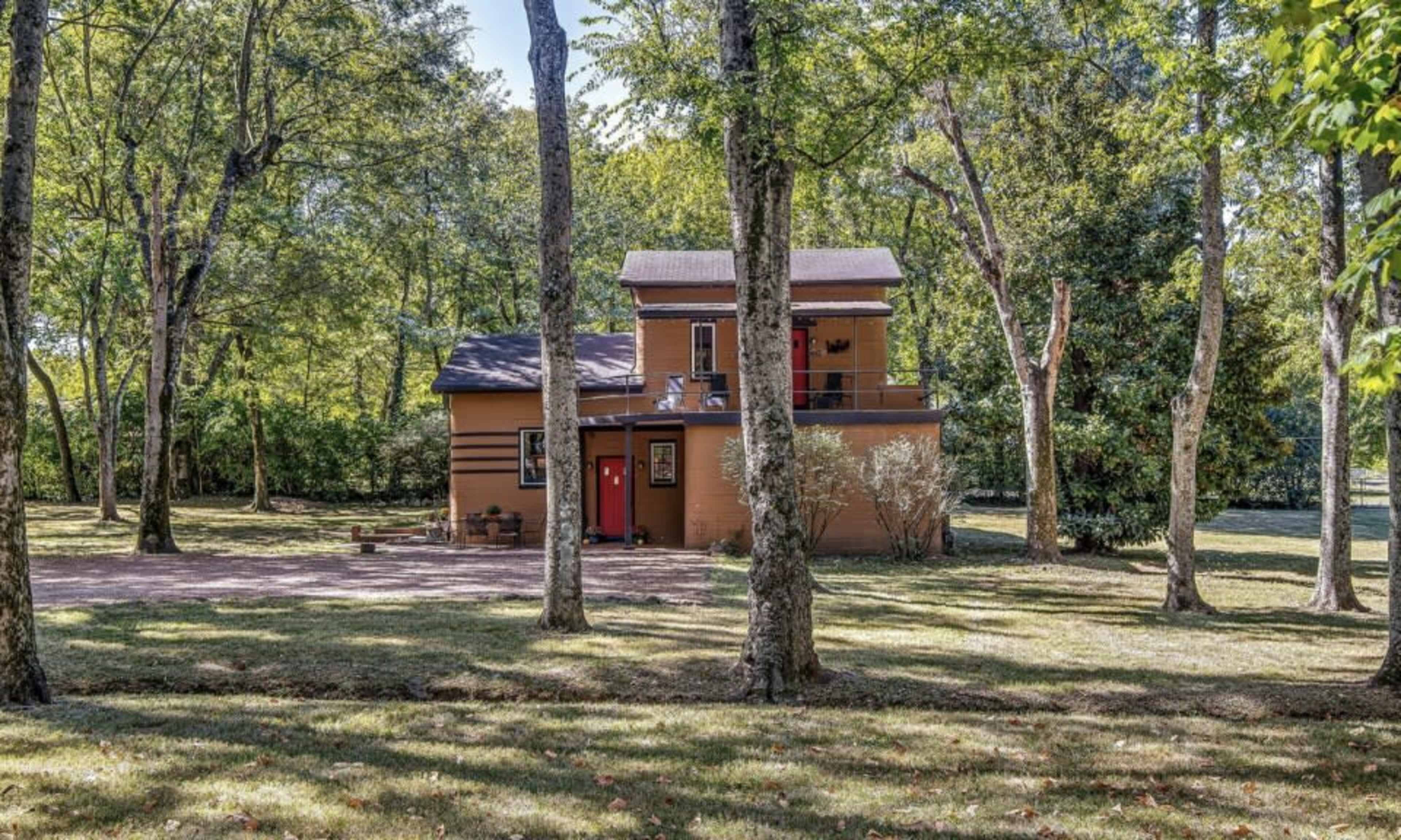 A two-story brown house is situated among tall trees in a grassy area.