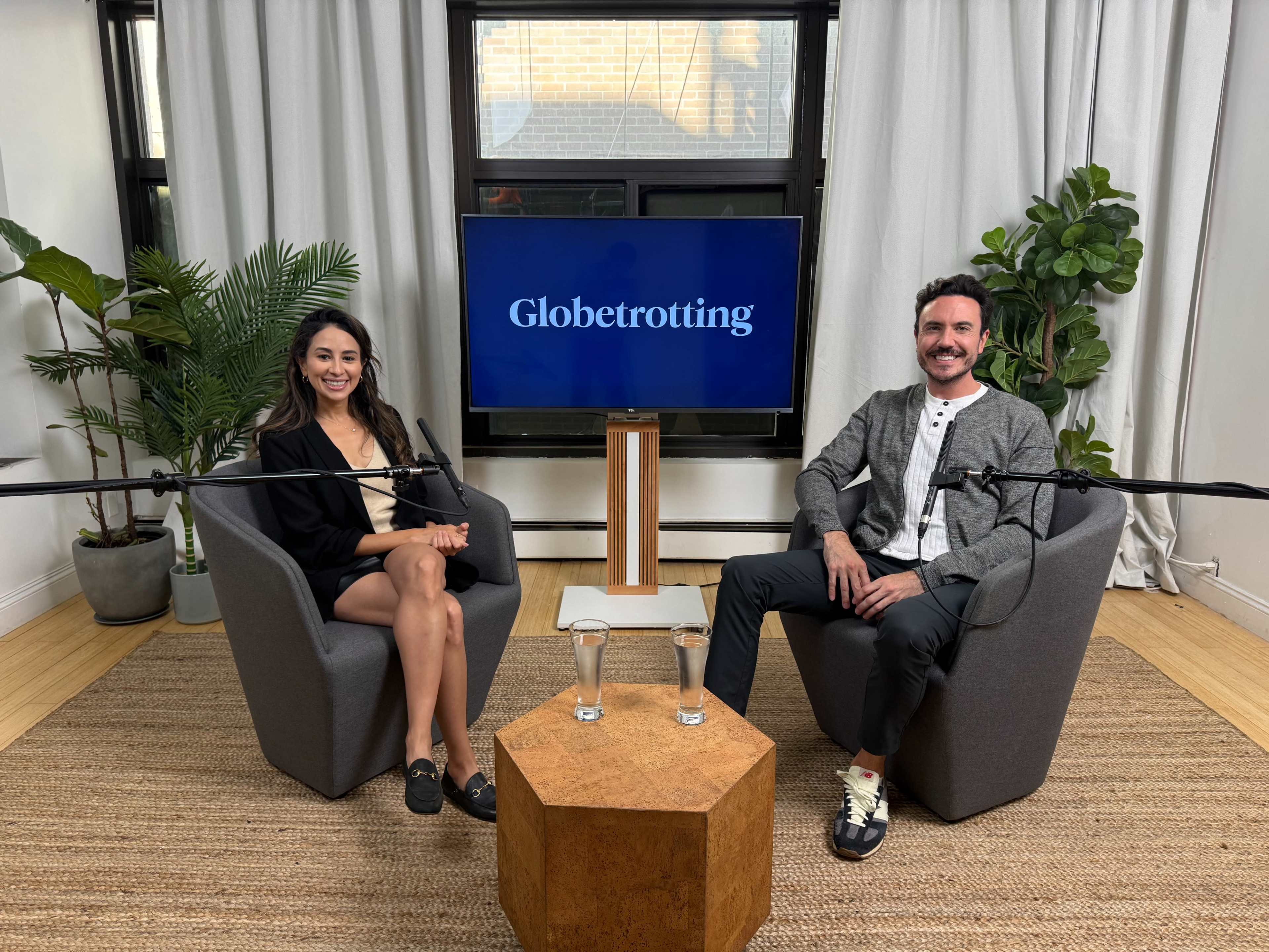 Two people are seated in a well-lit studio with a large screen displaying the word "Globetrotting" behind them, surrounded by plants and equipped with microphones.