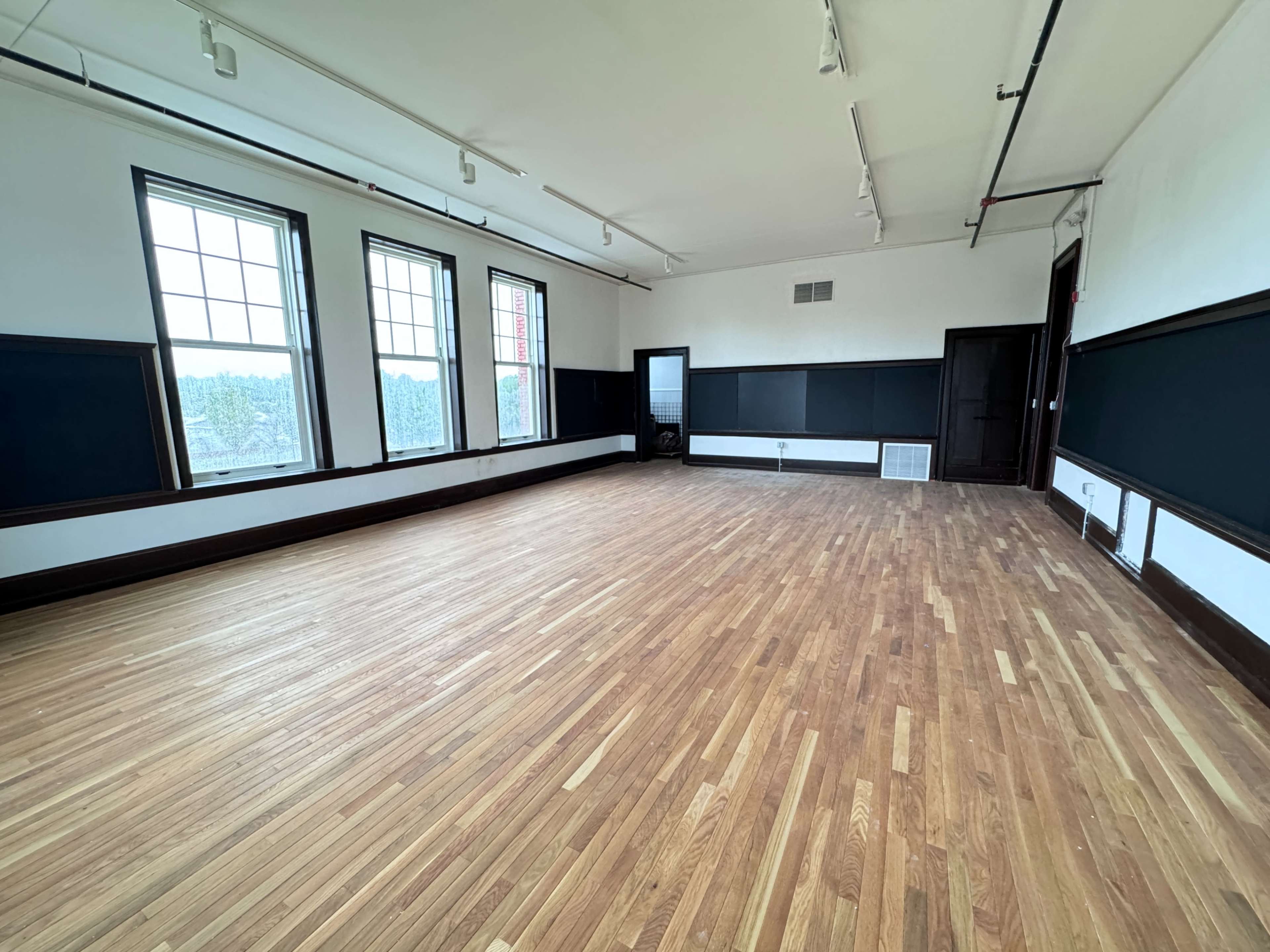 The image shows a spacious, empty room with hardwood floors and large windows along the walls.