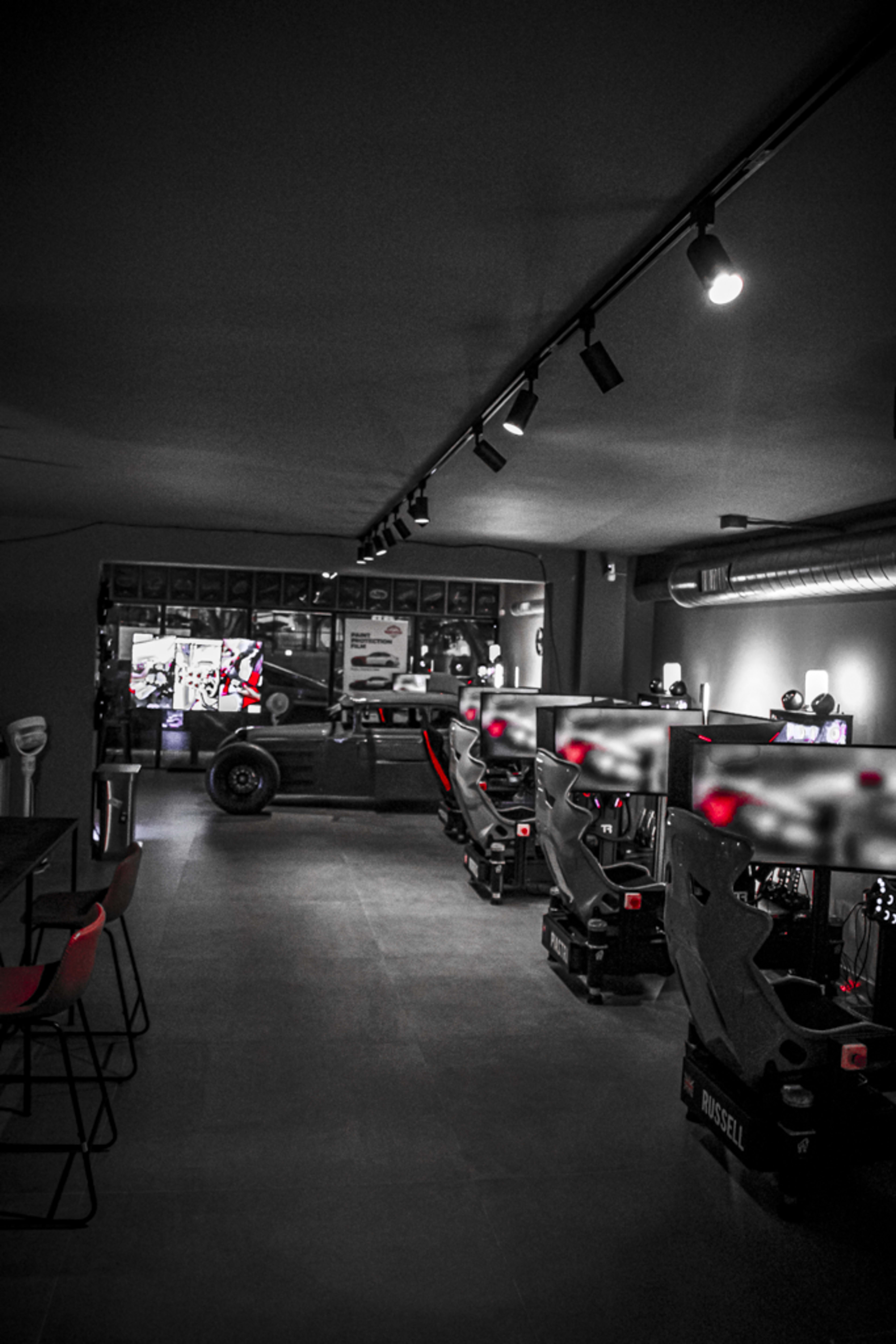 The image shows a modern gaming room with several gaming stations featuring black and red equipment, illuminated by overhead lights.