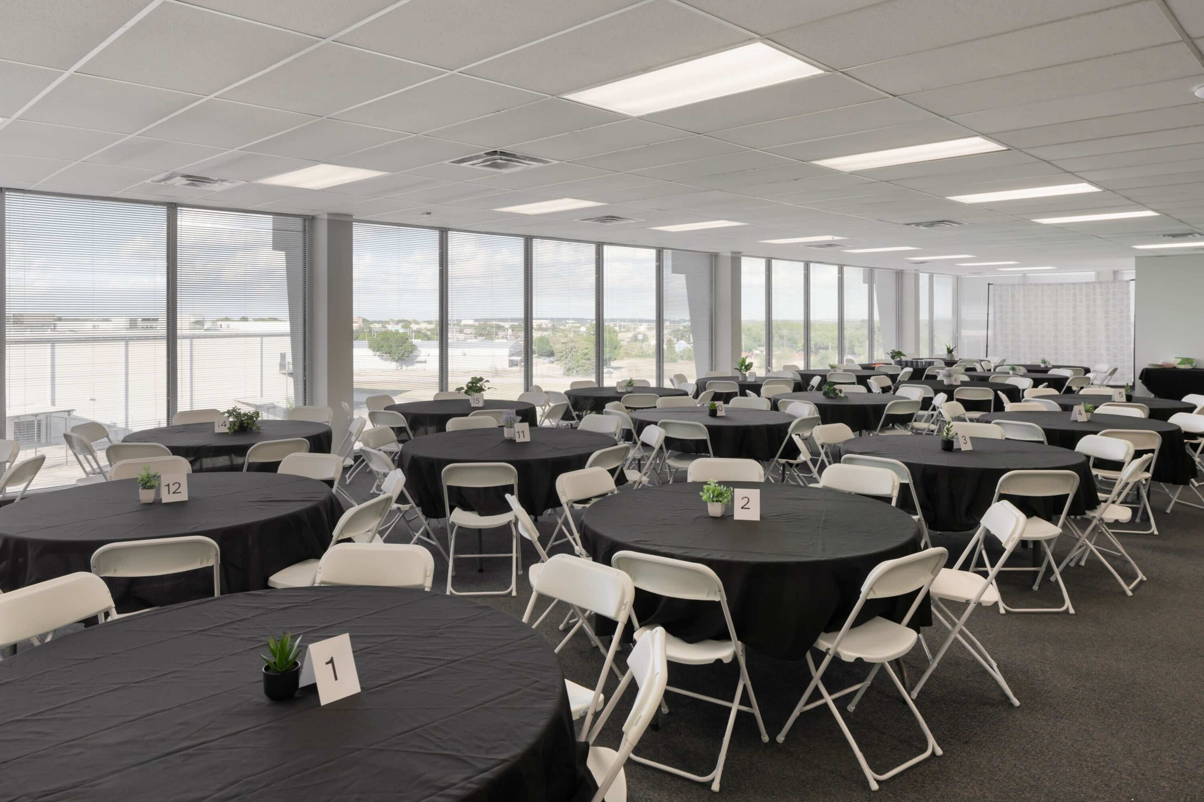 A spacious event room features multiple round tables covered with black tablecloths, each labeled with a number, and adorned with small green plants, along with large windows providing natural light.