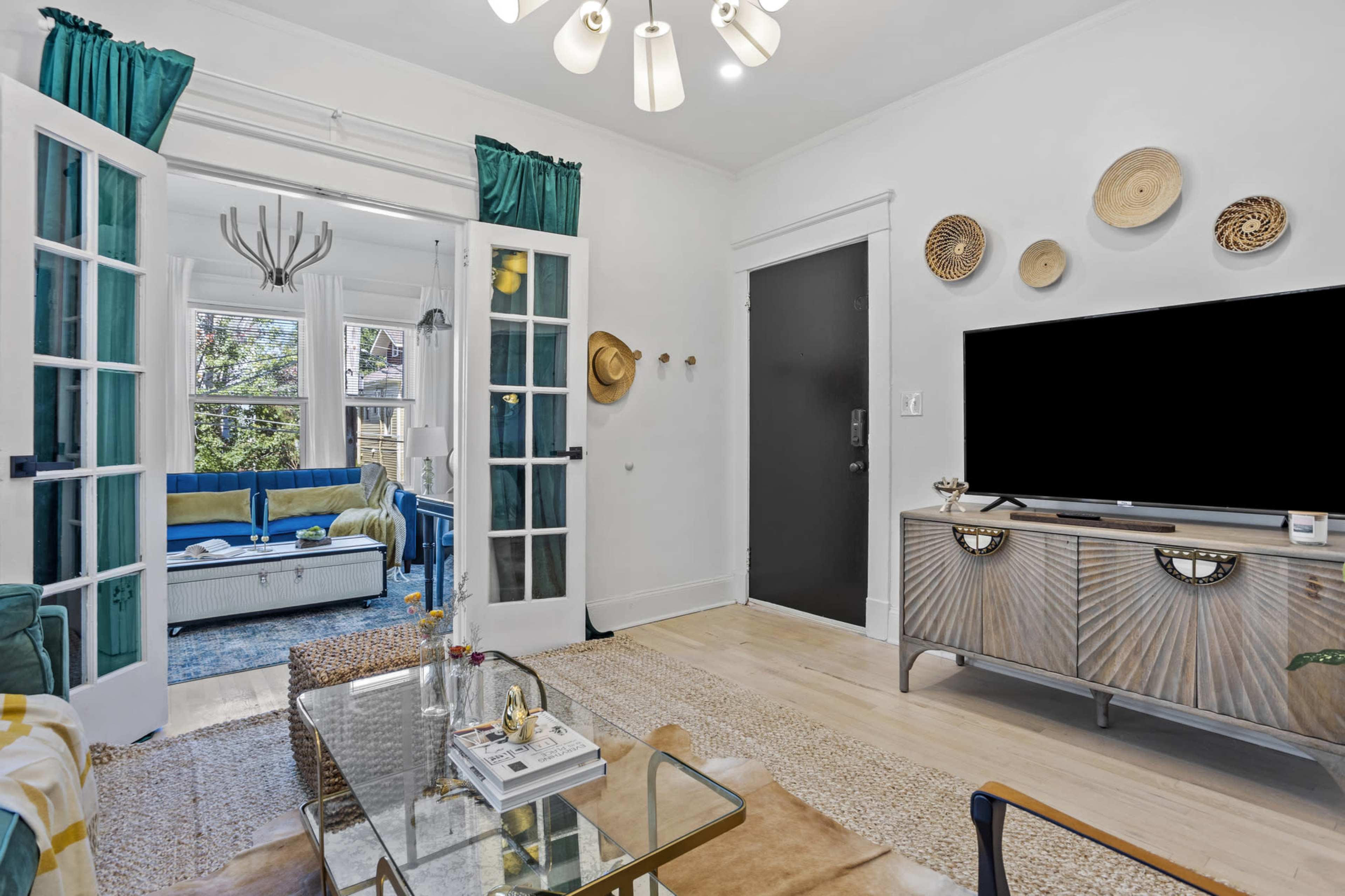 A modern living room features a glass coffee table, a media console, and French doors leading to a well-lit seating area.