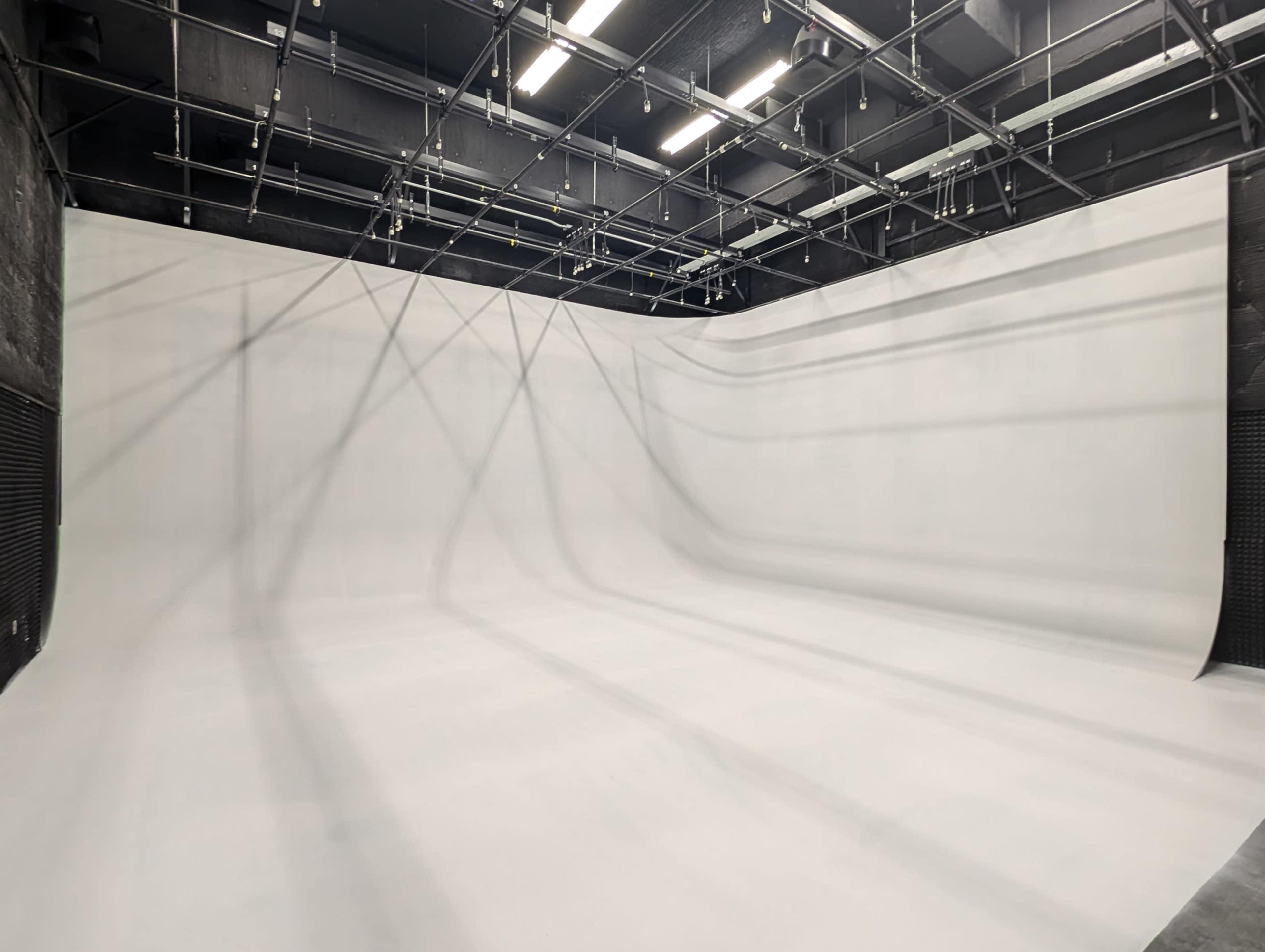 The image shows a large, empty studio space with a seamless white backdrop and overhead grid lights.