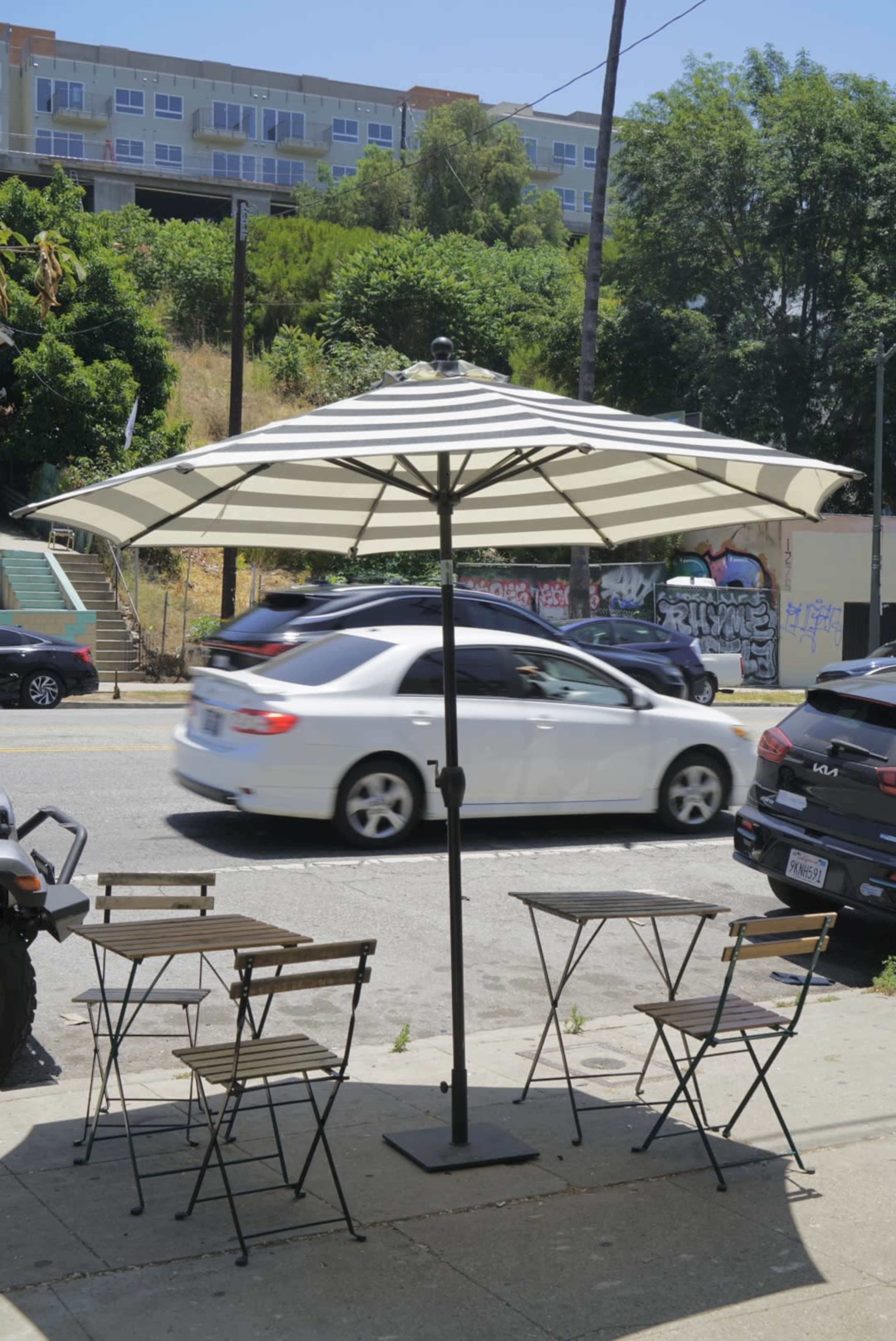 A large umbrella shades a set of metal tables and chairs on a sidewalk beside a busy street.