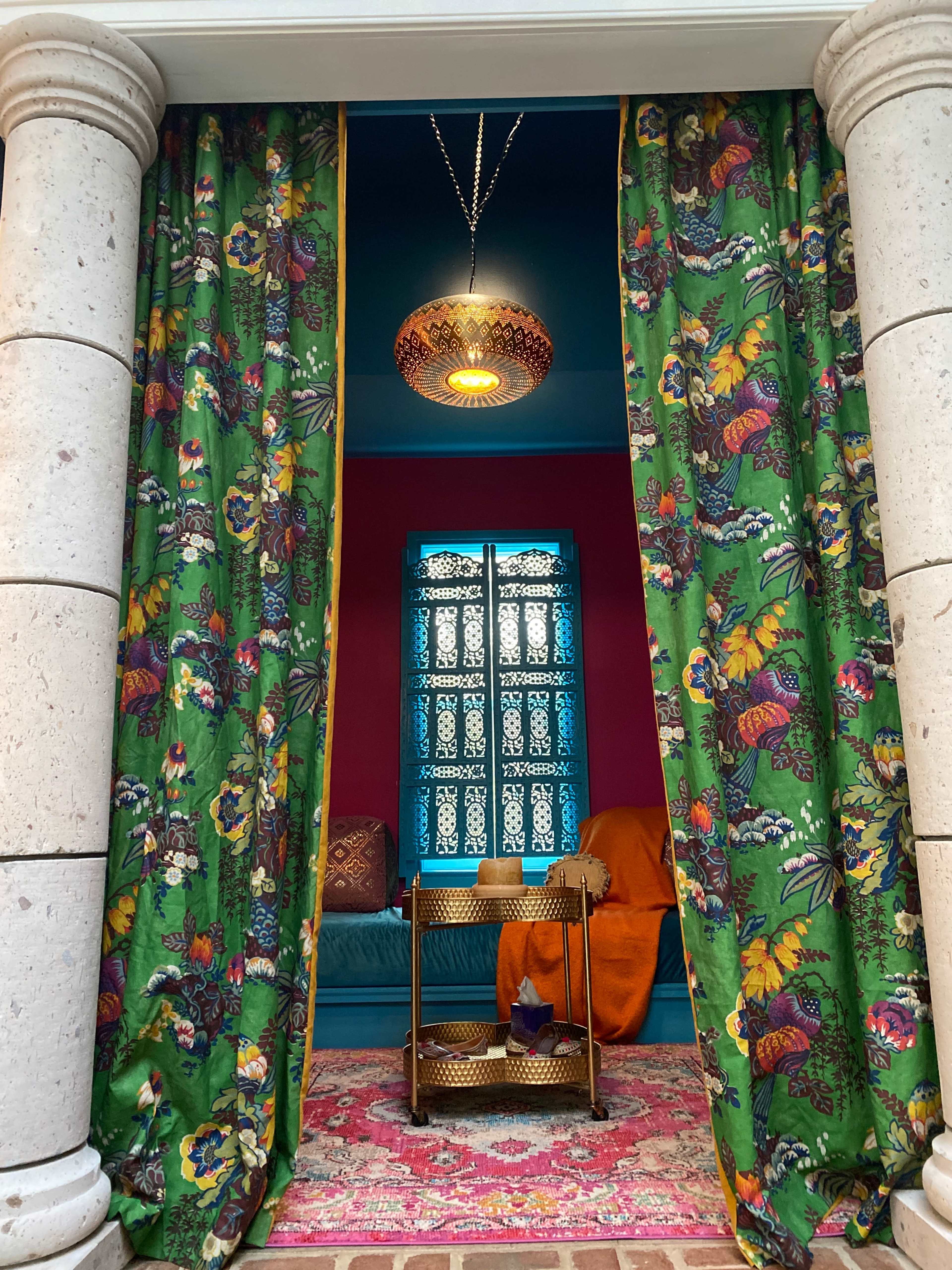 A colorful indoor space with floral curtains, a blue ceiling, a decorative light fixture, and a cozy seating area visible through a doorway.