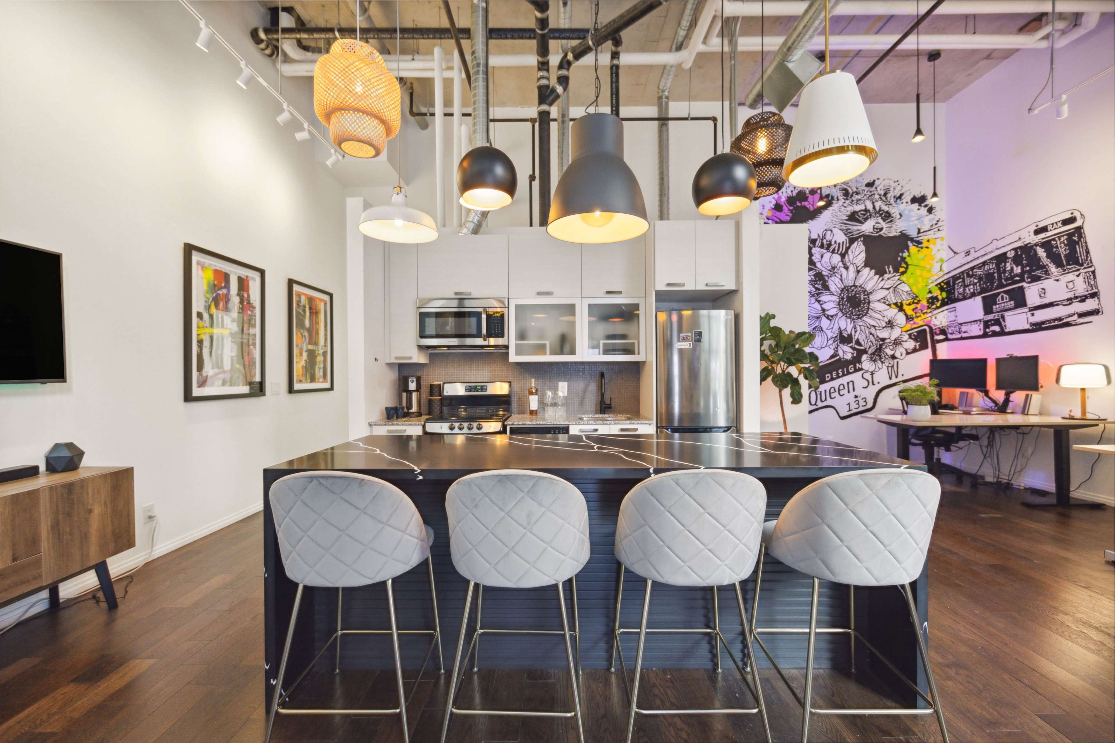 A modern kitchen features a central island with four barstools, pendant lighting, and a mix of appliances against a backdrop of vibrant wall art.