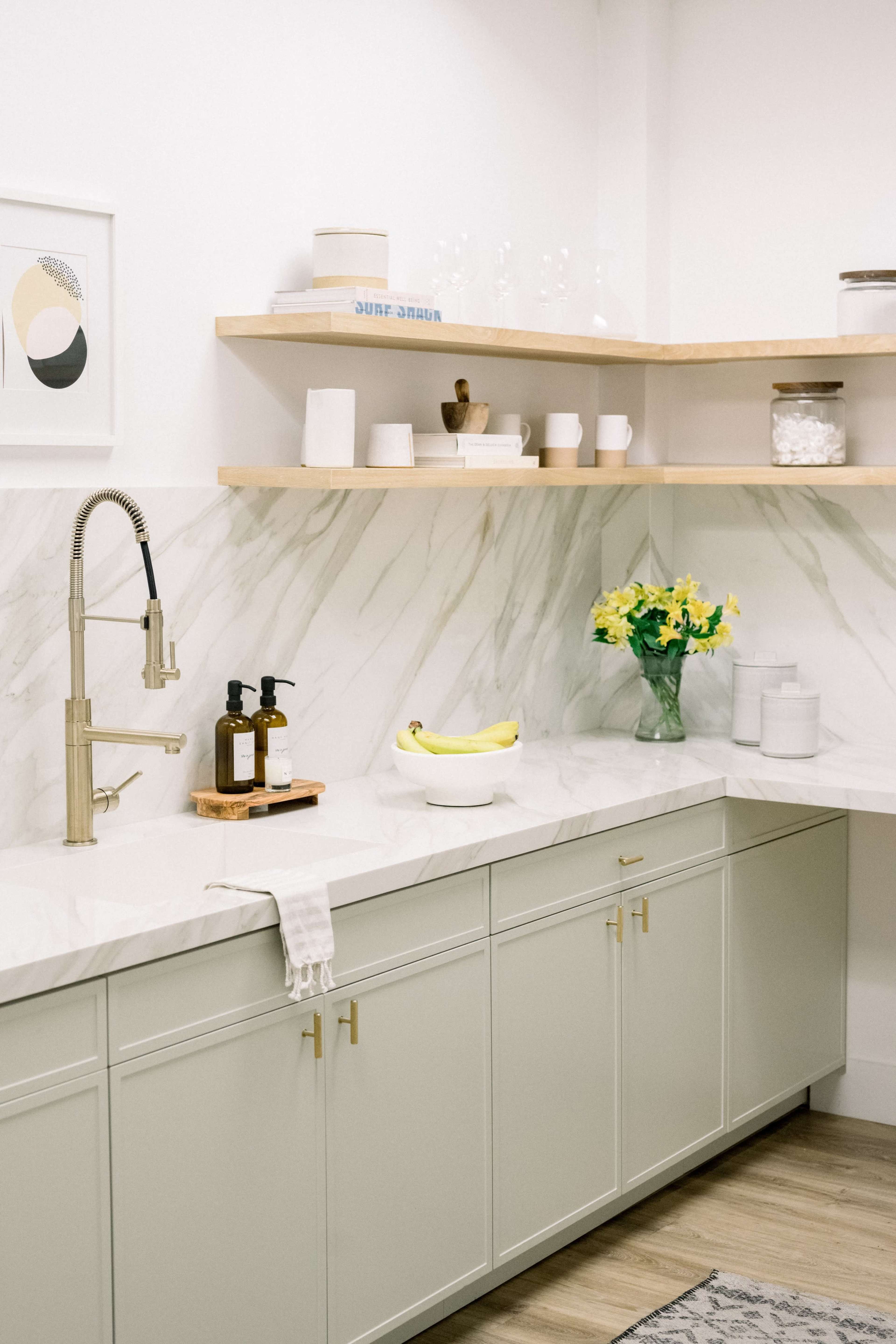 The image shows a modern kitchen with marble countertops, light green cabinetry, and open wooden shelves displaying various kitchen items and a bouquet of flowers.