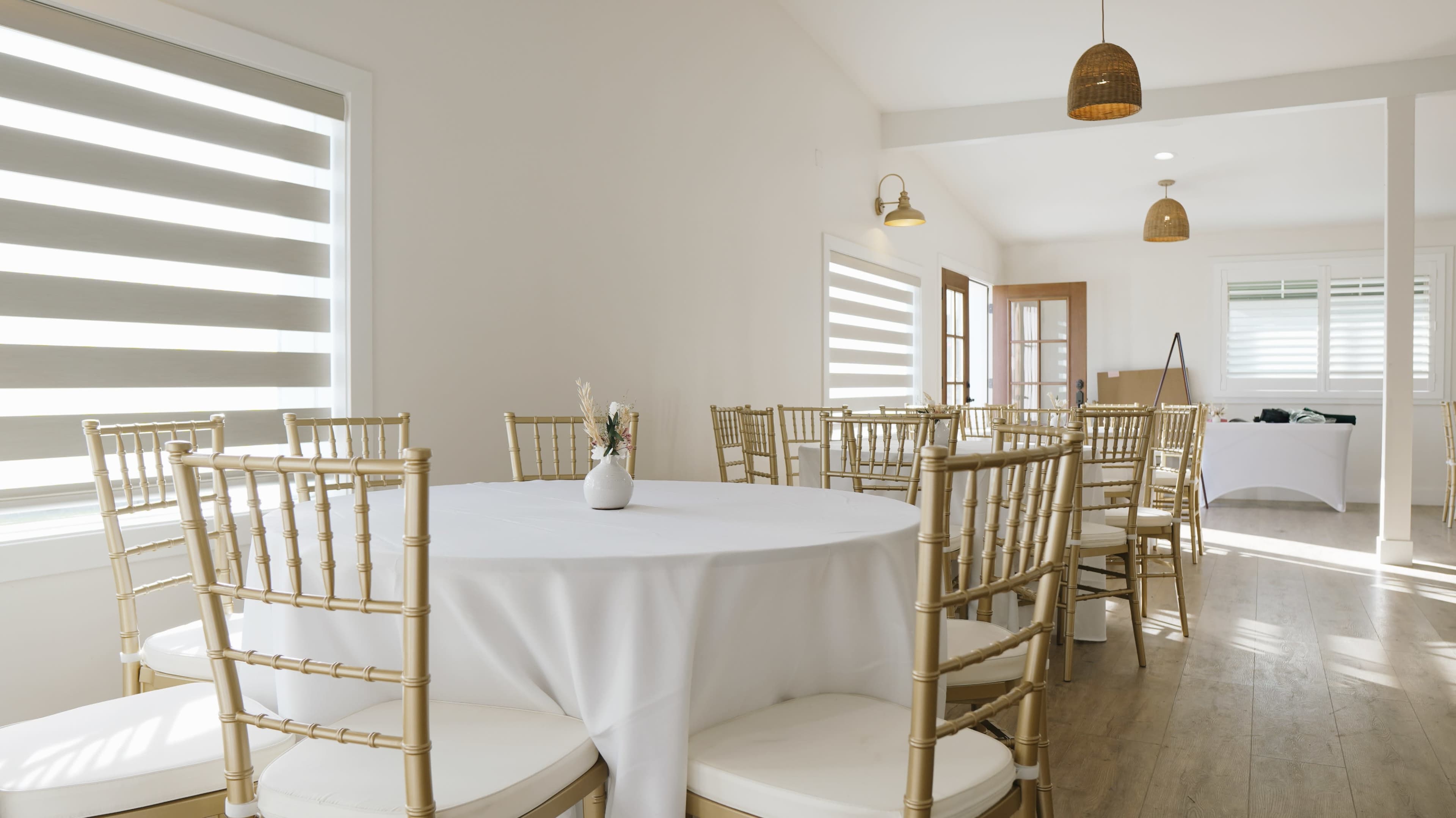 A set of elegantly arranged tables with gold chiavari chairs is positioned in a well-lit room featuring large windows with striped shades.