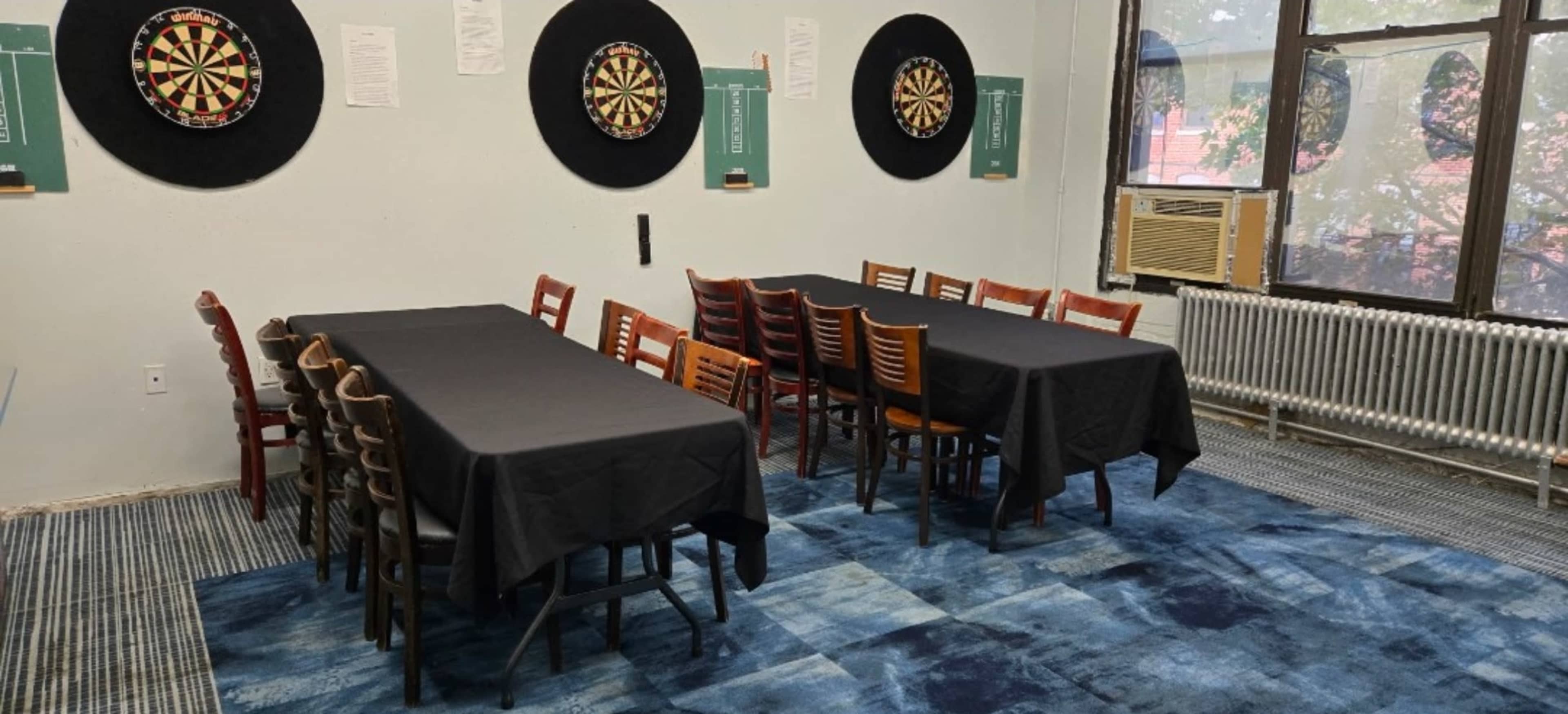 The image shows a room with two dartboards on the walls and several tables with chairs arranged around them.