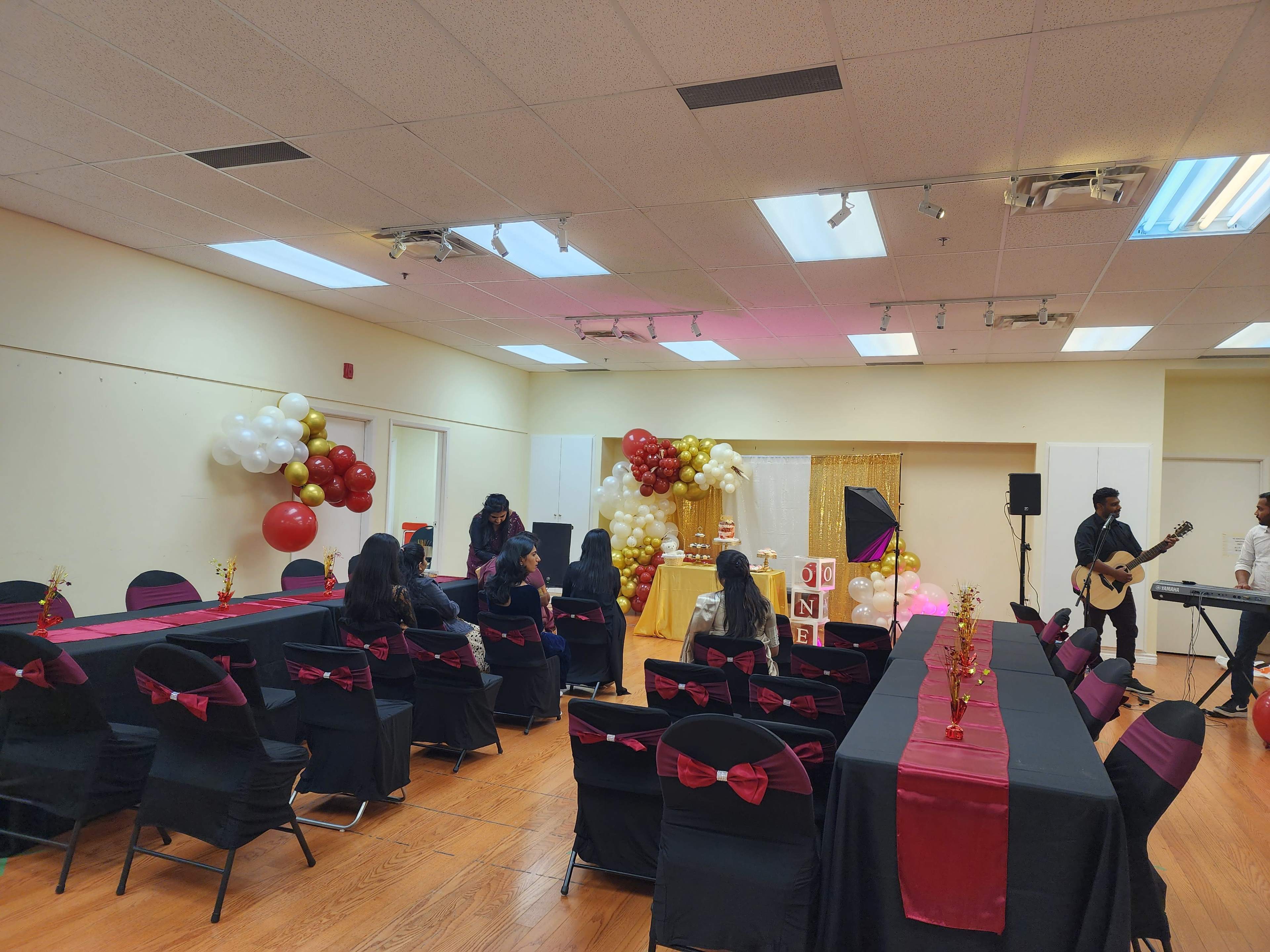 A decorated room with black tables and chairs featuring red bows, gold and white balloons, and a small stage set up for performers.