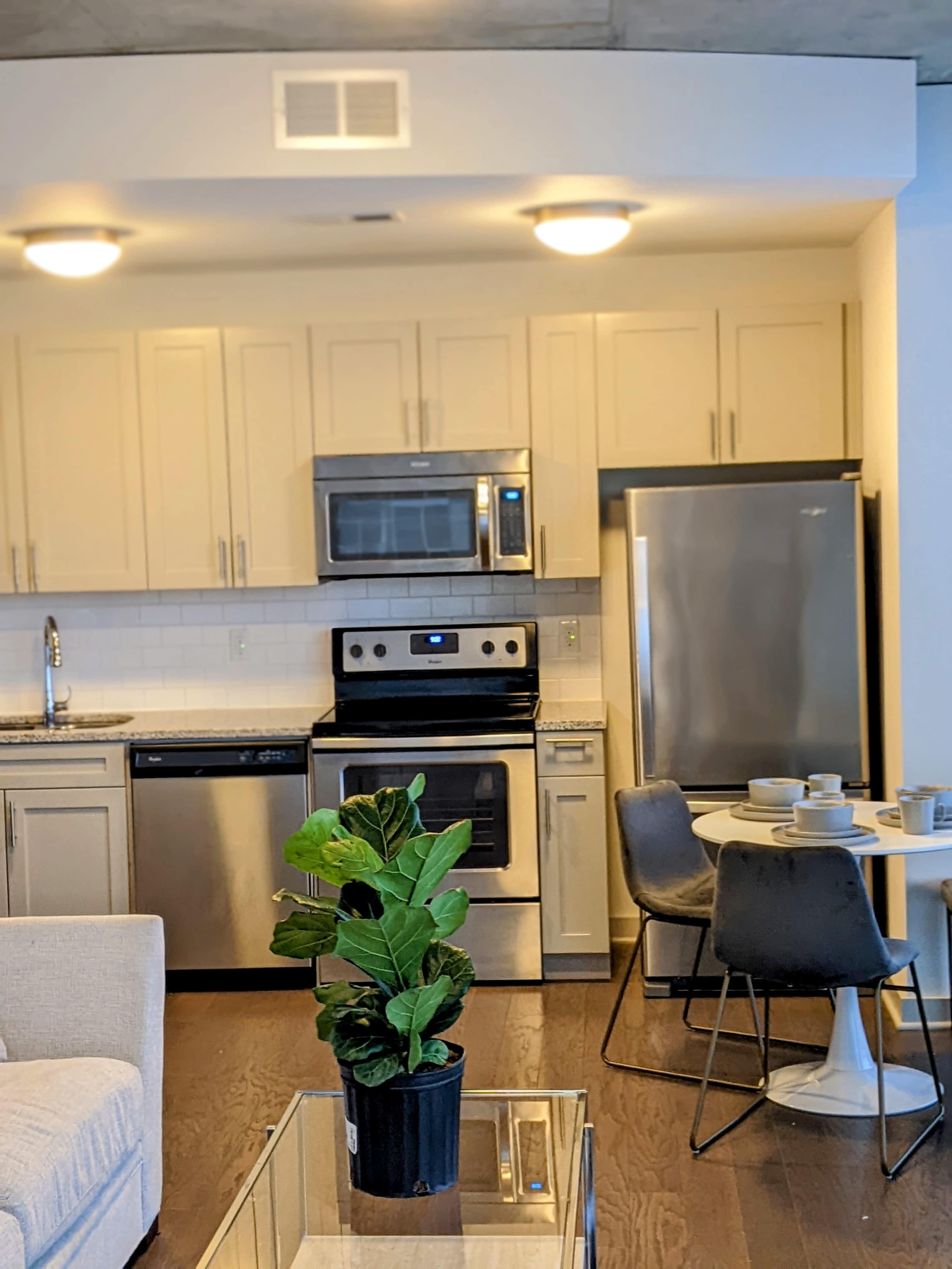 A modern kitchen features stainless steel appliances, light cabinetry, and a small dining table with chairs next to a couch in a living area.