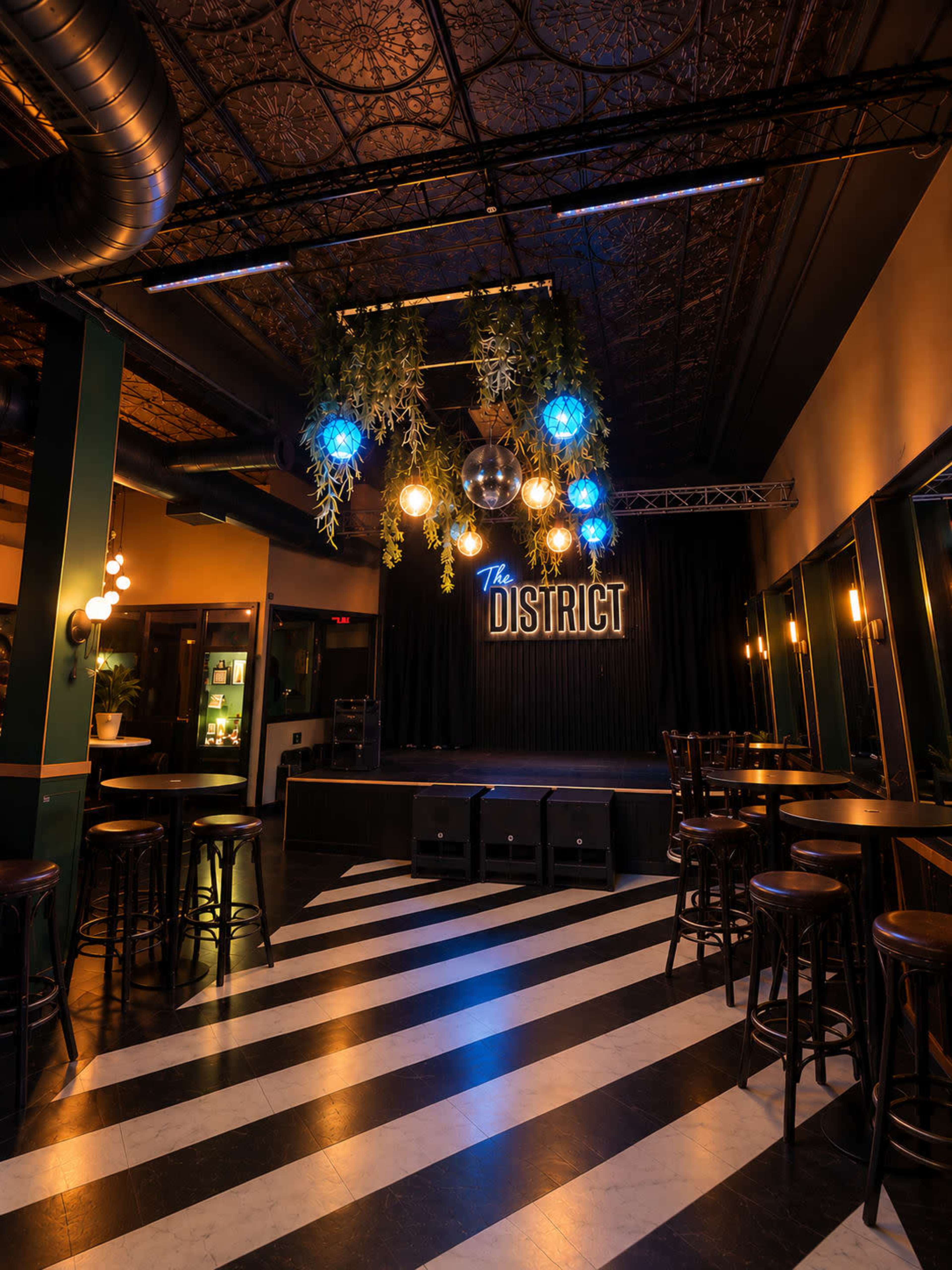 The image shows a dimly lit performance venue with a stage at one end, decorated with hanging lights and a sign that reads "The DISTRICT," and black-and-white striped flooring leading toward the stage.