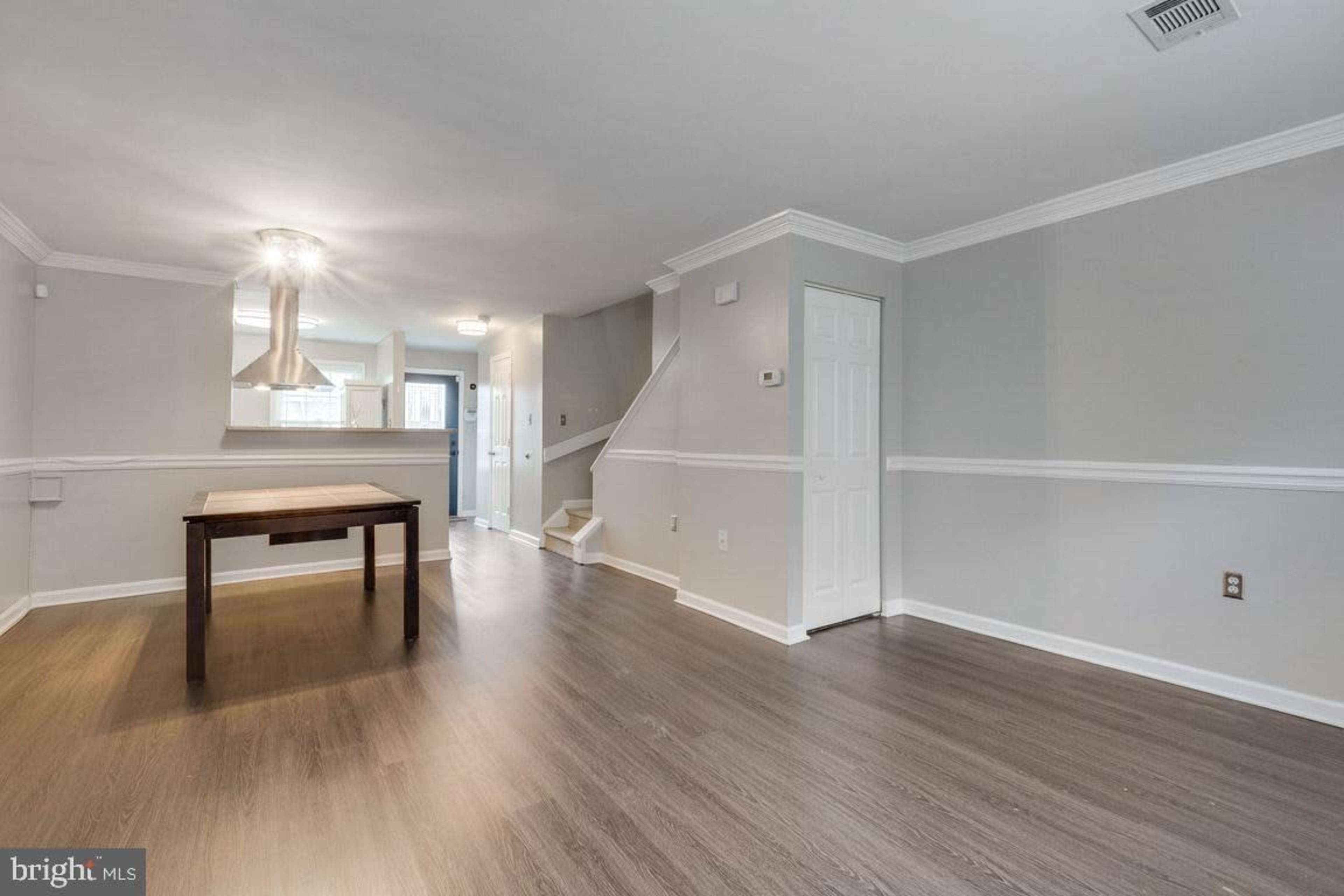 The image shows a modern, empty living space featuring a hardwood floor and a table, with a staircase in the background and a kitchen area visible through an open doorway.