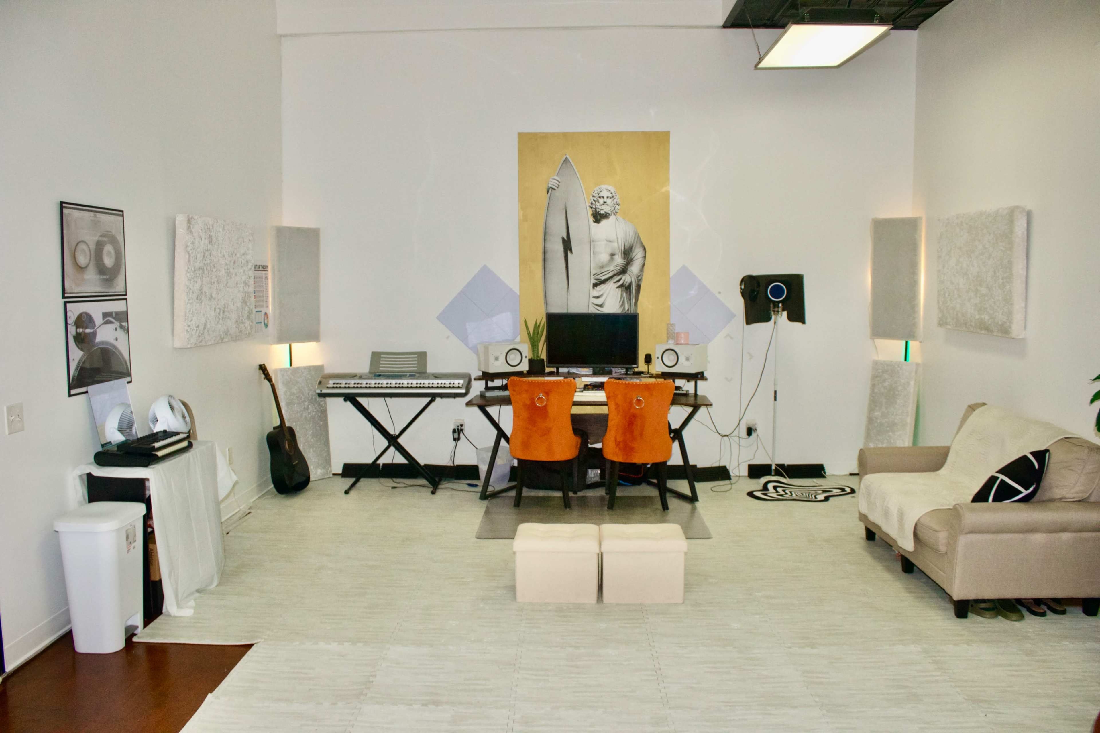 The image shows a modern music studio featuring a keyboard, two desks with chairs, artwork on the walls, and a sofa, all arranged in a spacious, well-lit room.
