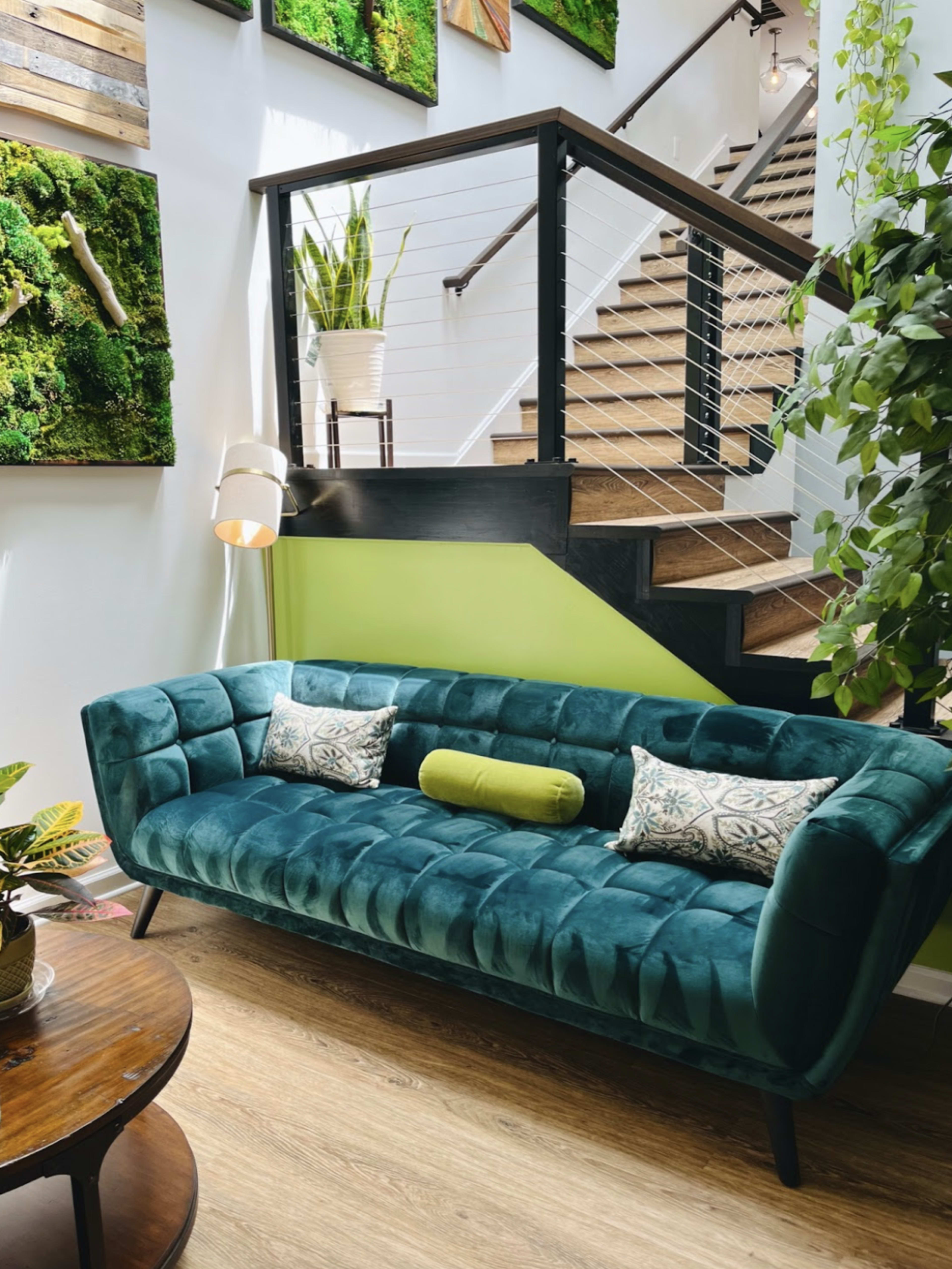 A green velvet sofa with decorative pillows is positioned near a staircase, surrounded by plants and wall-mounted greenery.