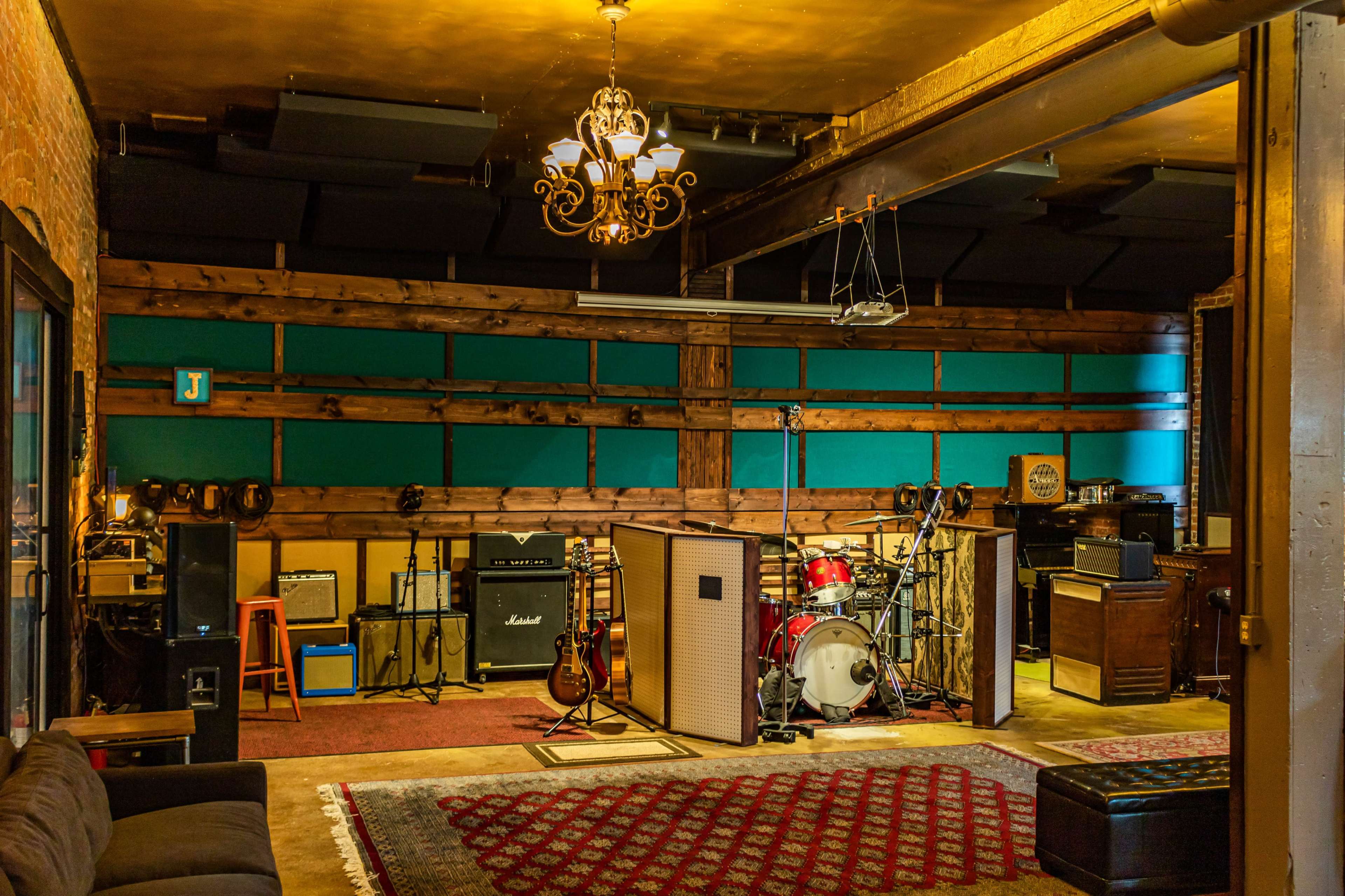 A music rehearsal space featuring guitars, amplifiers, and a drum set against a backdrop of wooden walls and teal panels.