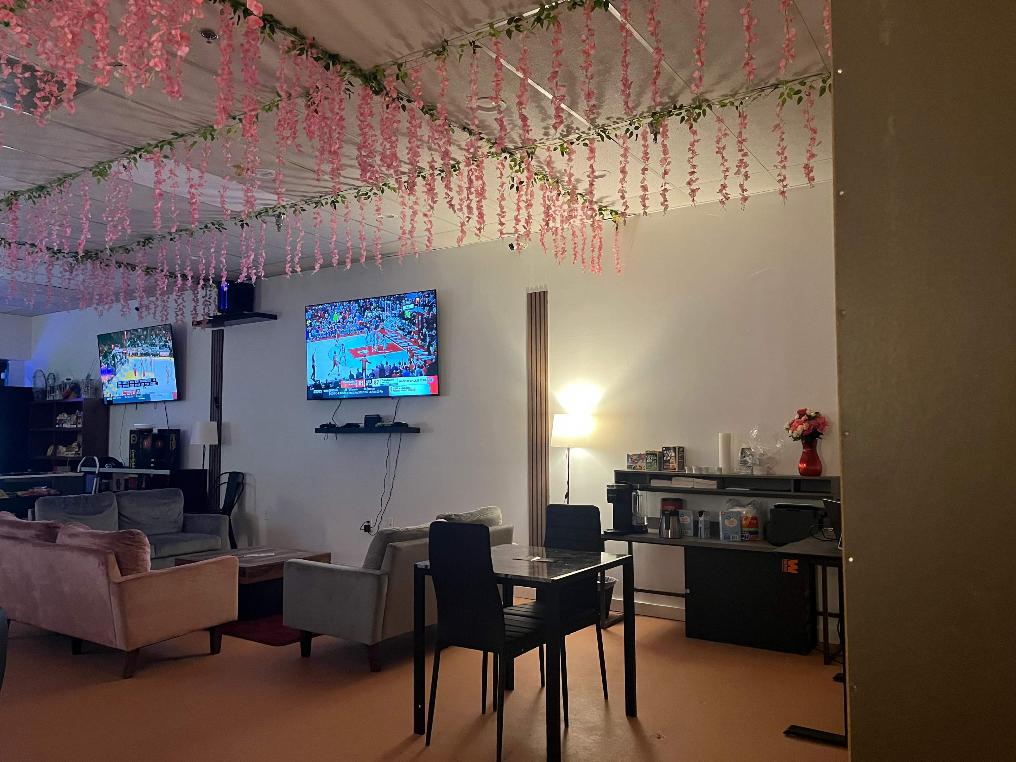 The image shows a cozy interior space with two televisions mounted on the wall, surrounded by pink decorations hanging from the ceiling, and furnished with a dining table, chairs, and a couch set.