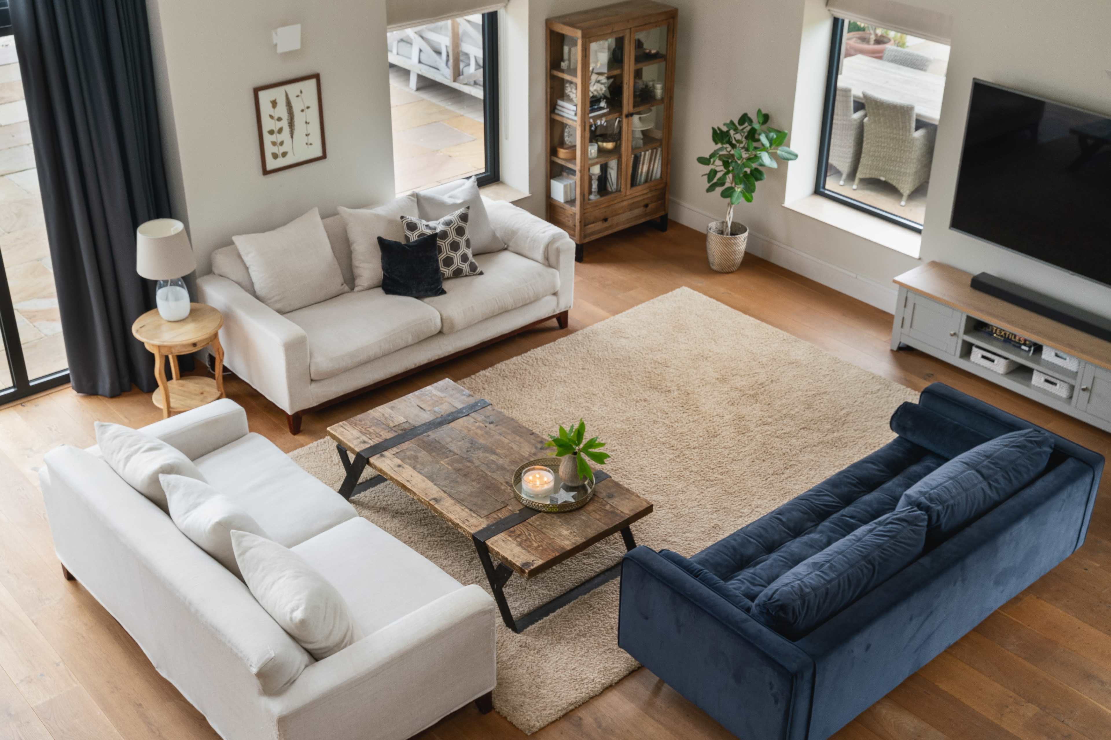 The image shows a cozy living room with three sofas arranged around a wooden coffee table on a large area rug, flanked by windows and a plant.