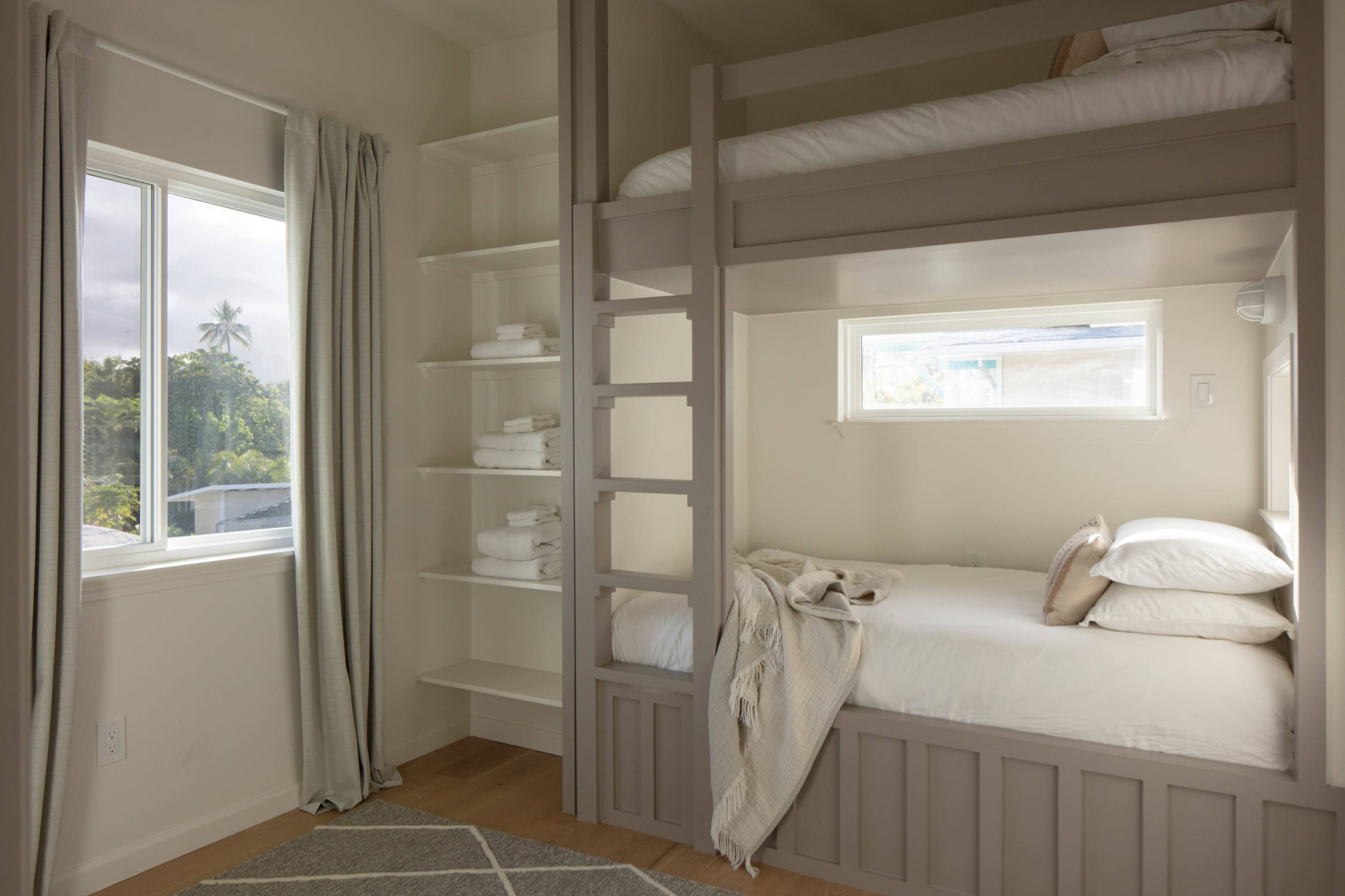 The image shows a small, well-lit bedroom with a bunk bed, a neatly made lower bed, and a window with light filtering through sheer curtains.