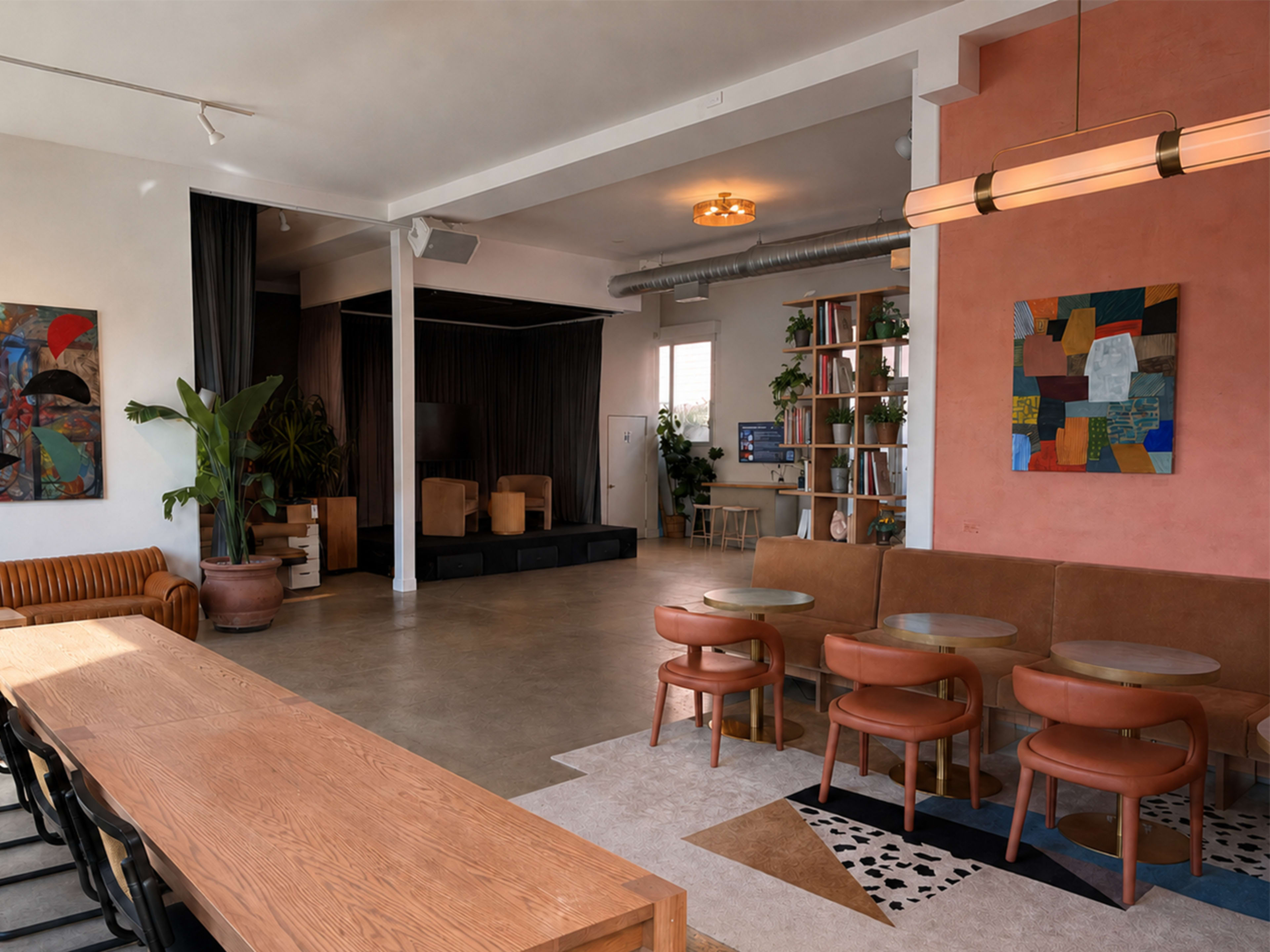 The image shows a spacious interior of a modern café or community space featuring a mix of seating arrangements, a stage area, and decorative plants.