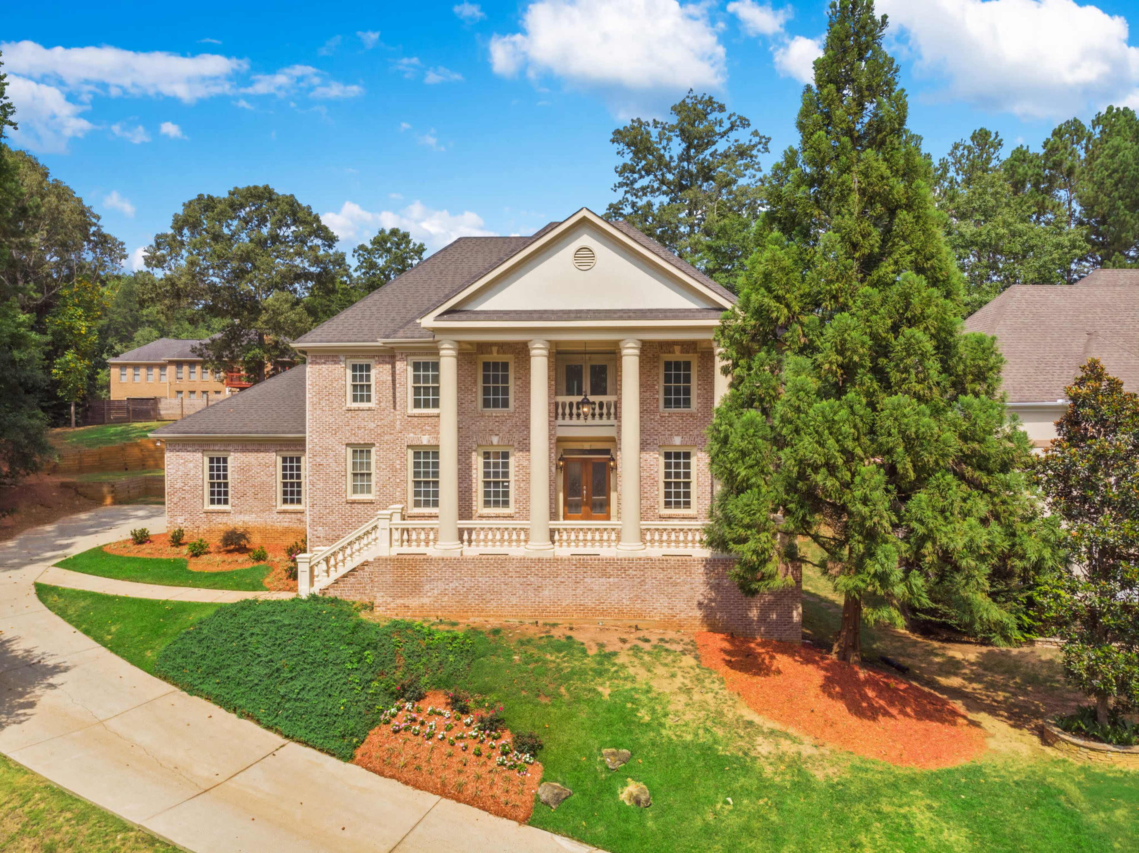 The image shows a large, two-story brick house with columns, surrounded by landscaped greenery and a paved driveway.